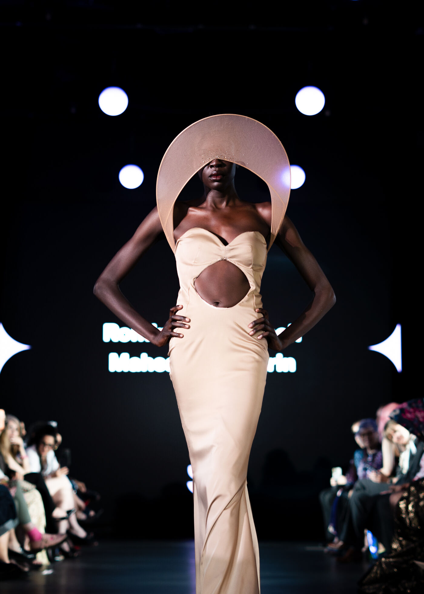 A woman in a long gown and a large, circular hat walks down a runway in front of an audience, with bright lights in the background.