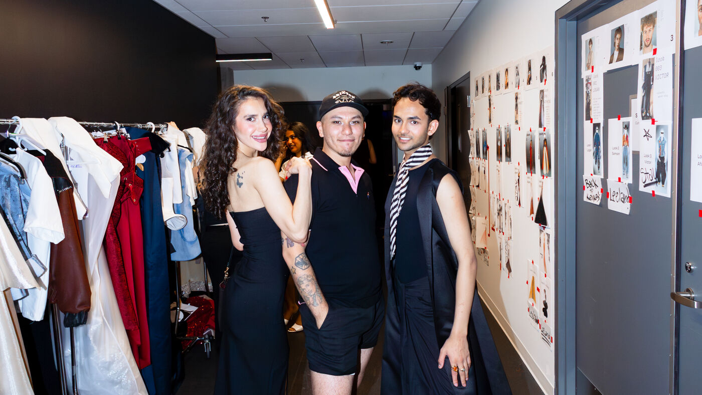 Three people stand in a hallway with clothes racks on the left and images on the wall to the right. The person on the left wears a black dress, the middle person wears a cap and black polo shirt, and the person on the right wears a black outfit.