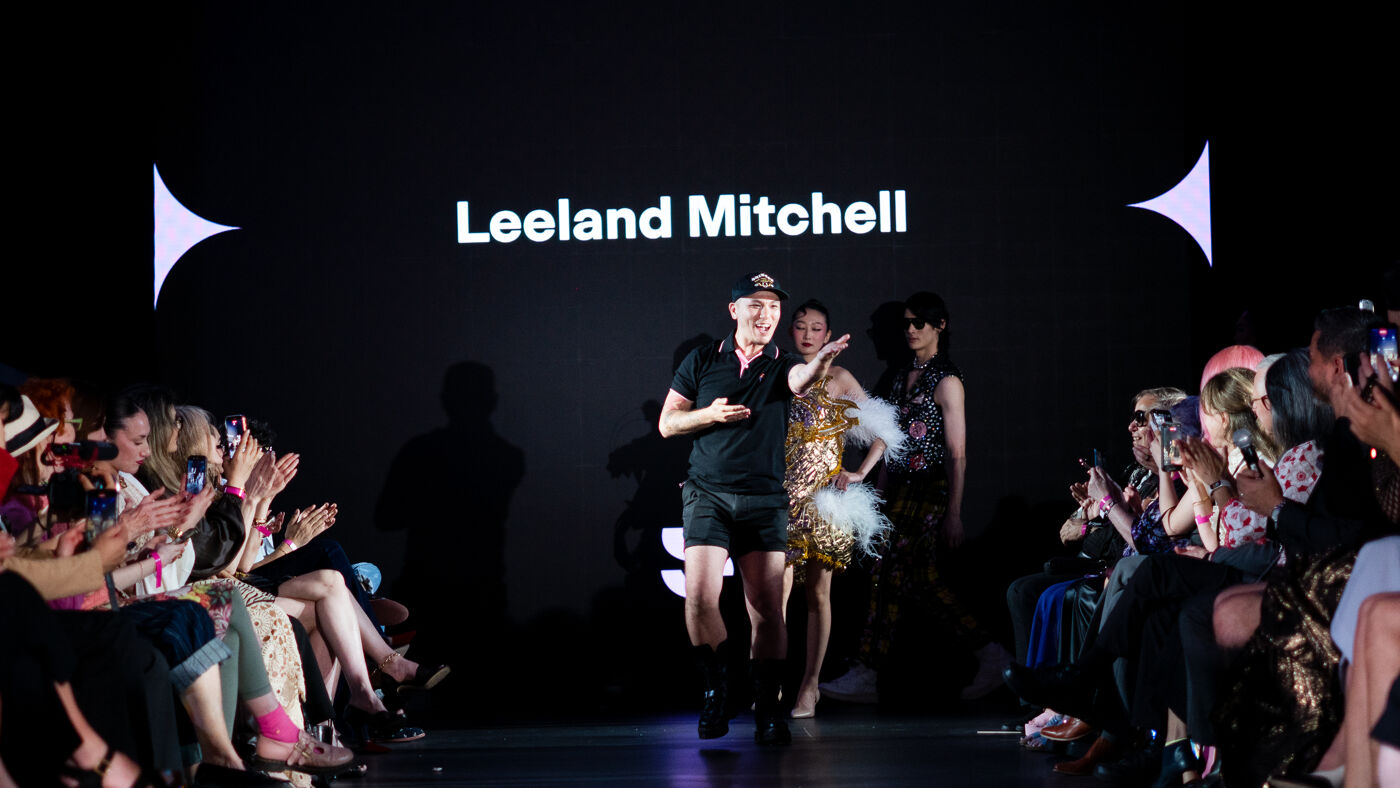 Fashion designer Leeland Mitchell walks the runway with models as the audience applauds. The screen displays his name.