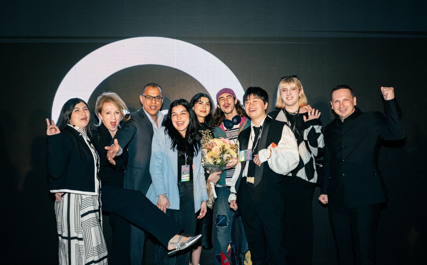 A group of eleven people, predominantly women, pose for a photo, some holding a bouquet of flowers, with a large white circle behind them.