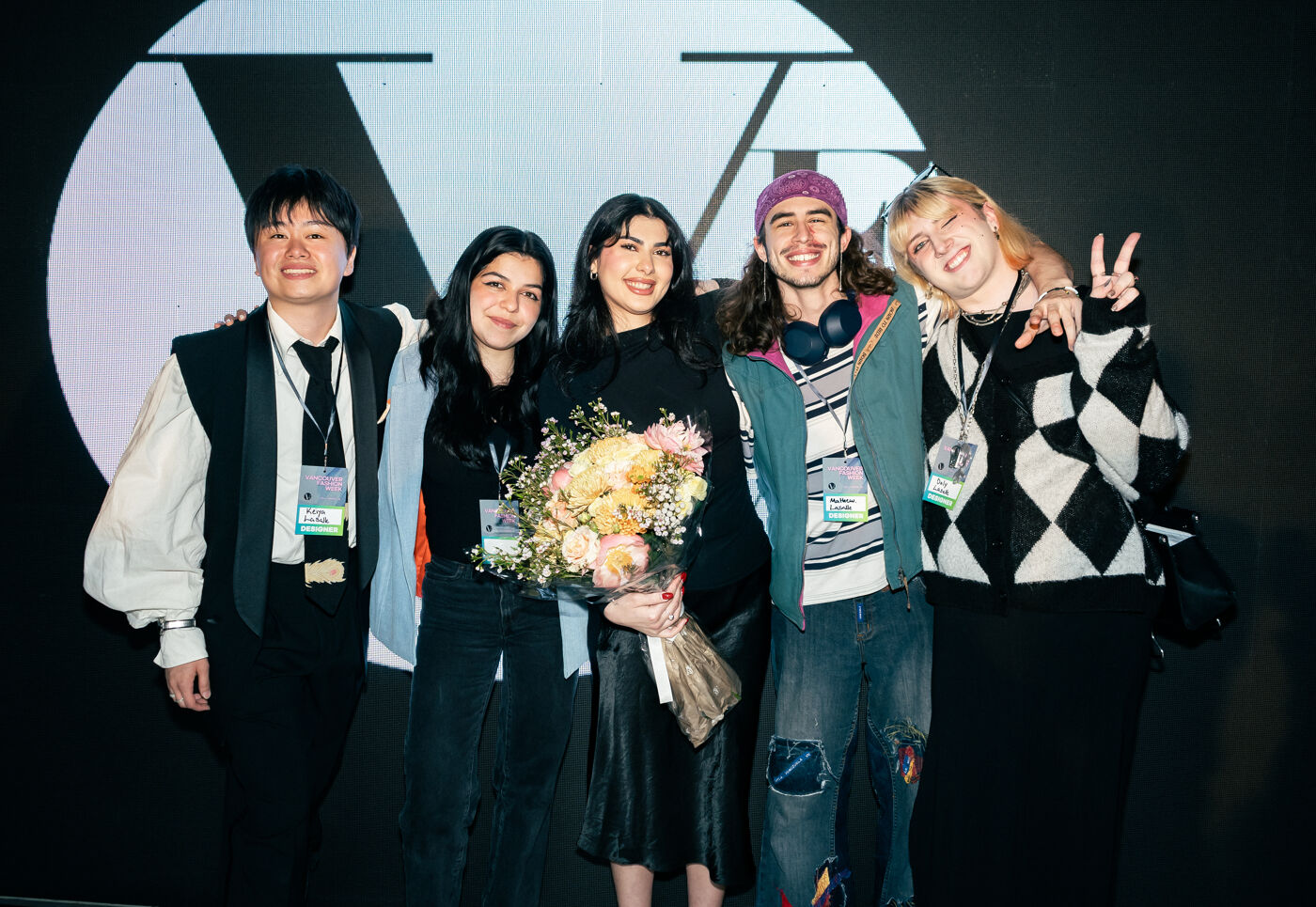 Five young adults, two men and three women, stand closely together, smiling, holding a bouquet of flowers.
