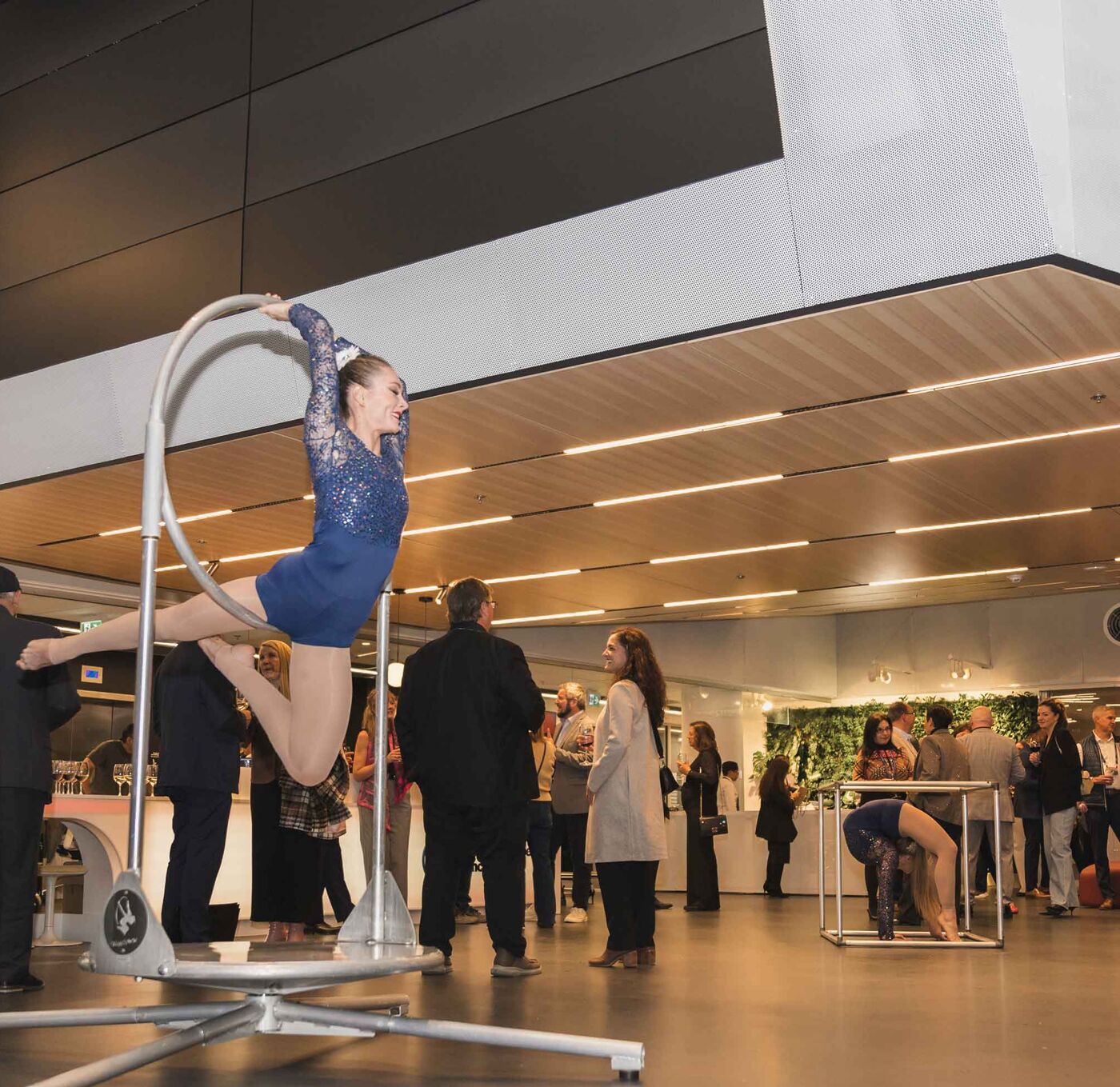 A young woman in a blue leotard performs an acrobatic move on a circular apparatus, with spectators watching in the background.