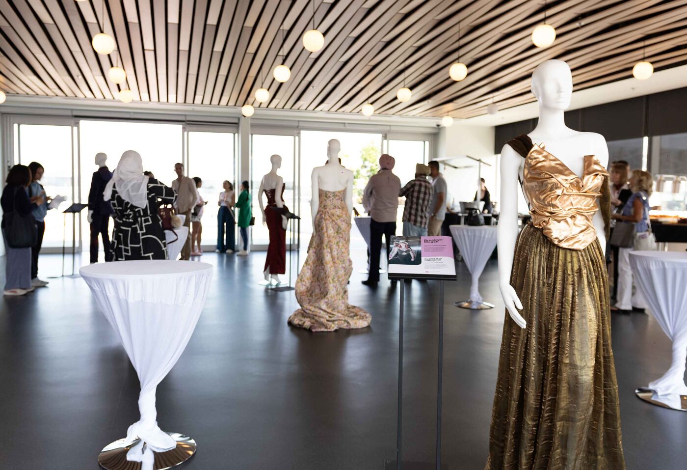 This image shows a fashion exhibition with several mannequins displaying elegant gowns. Attendees are observing the displays in a well-lit, modern venue with unique architectural ceiling details.