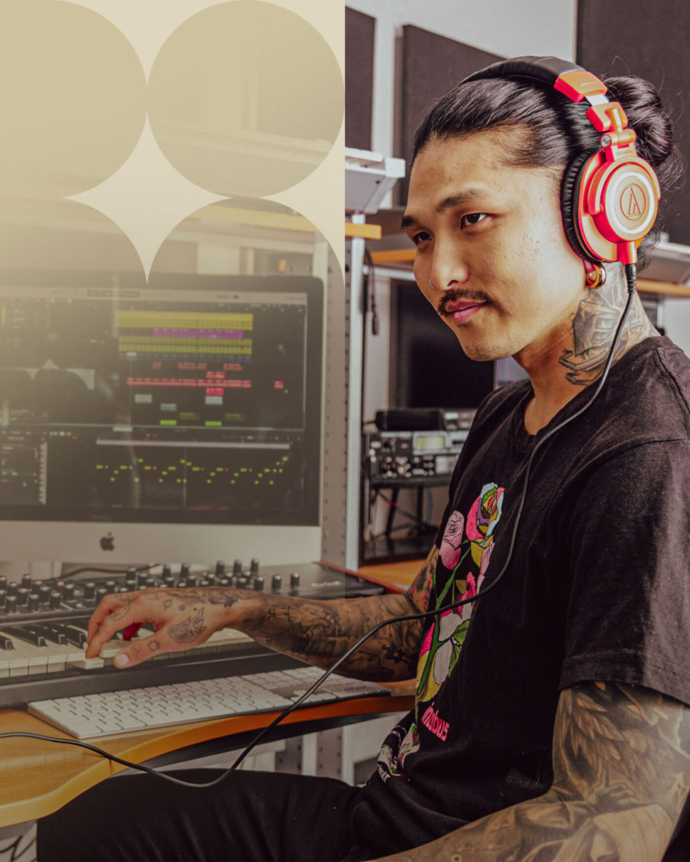 A tattooed man wearing headphones works in a recording studio, focused on a computer displaying music production software.