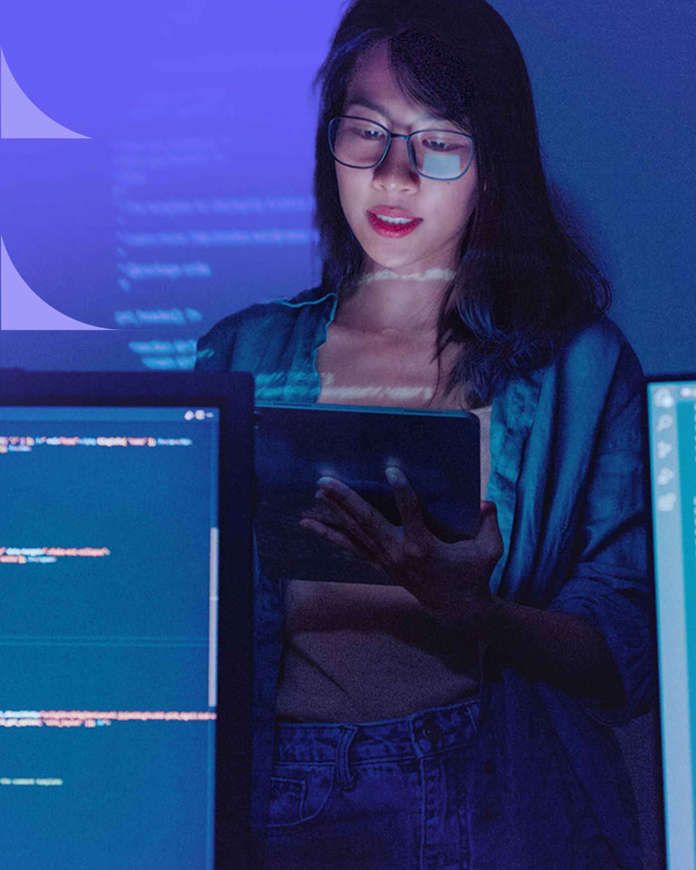 A woman wearing glasses looks at a tablet while surrounded by screens displaying code, illuminated by the blue light from the monitors.
