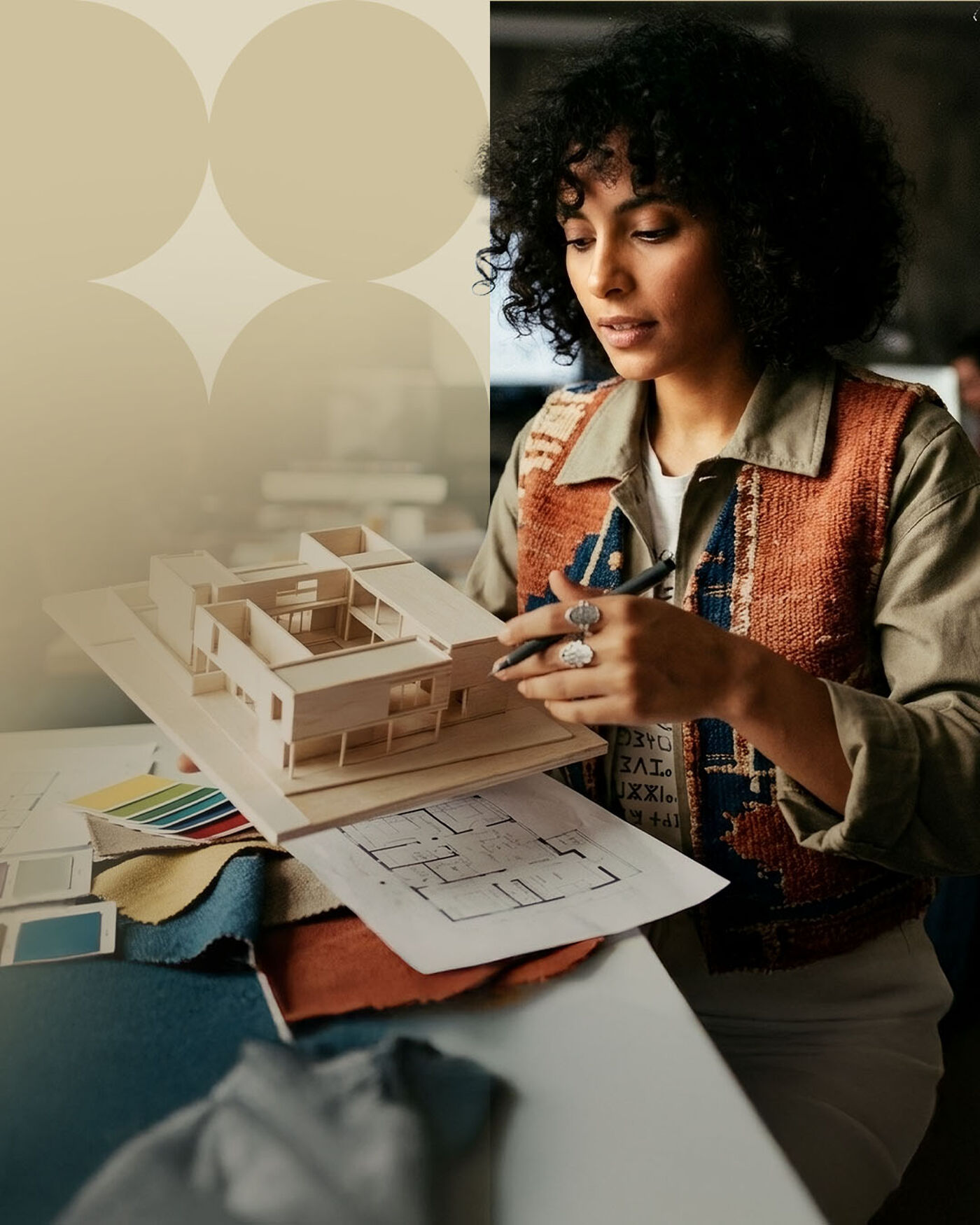A woman with curly black hair, wearing a colorful vest over a blouse, holds a pencil and examines a wooden architectural model on a desk with blueprints and fabric swatches.
