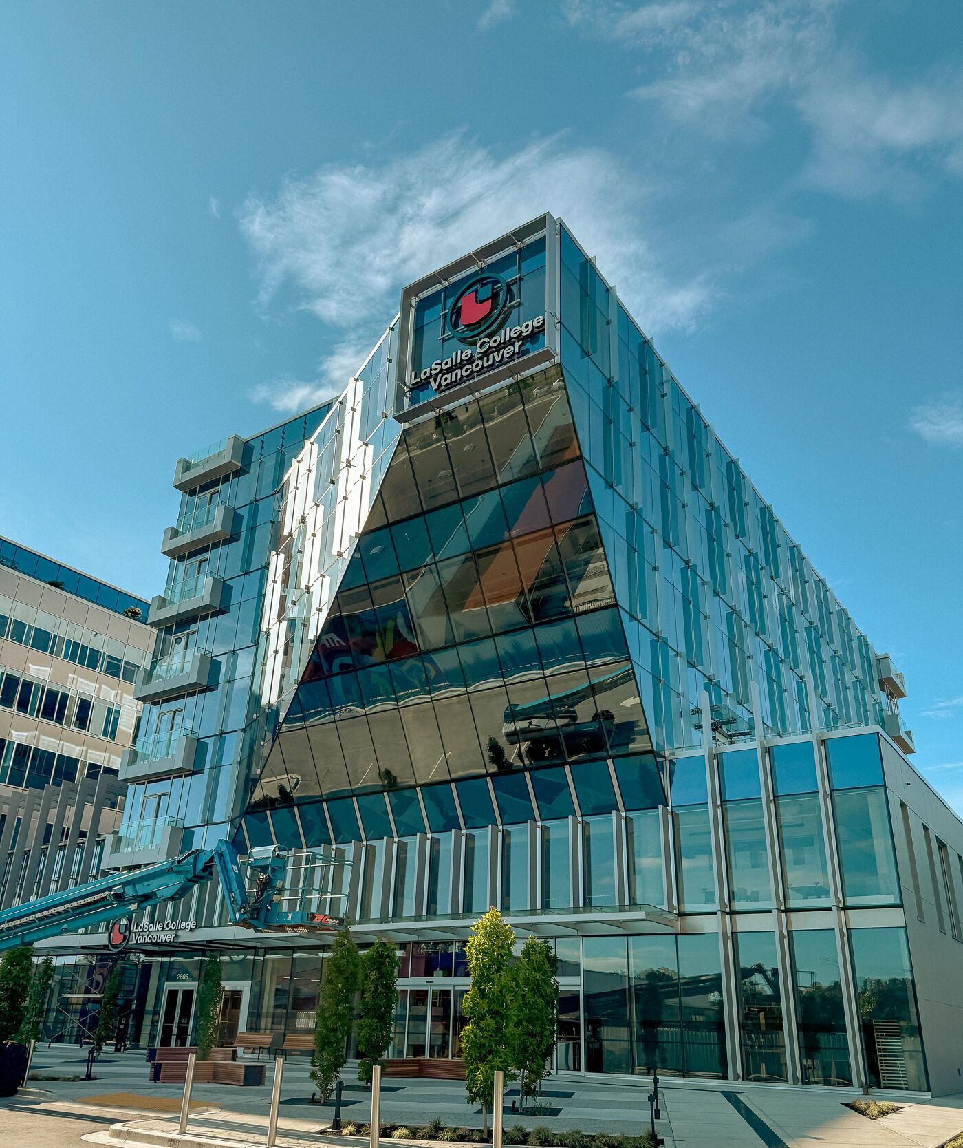 The image shows a modern, multi-story building with a unique architectural design featuring a prominent glass facade. The building appears to be a college or university, with a logo visible on the upper section. Landscaping is present at the base of the building.