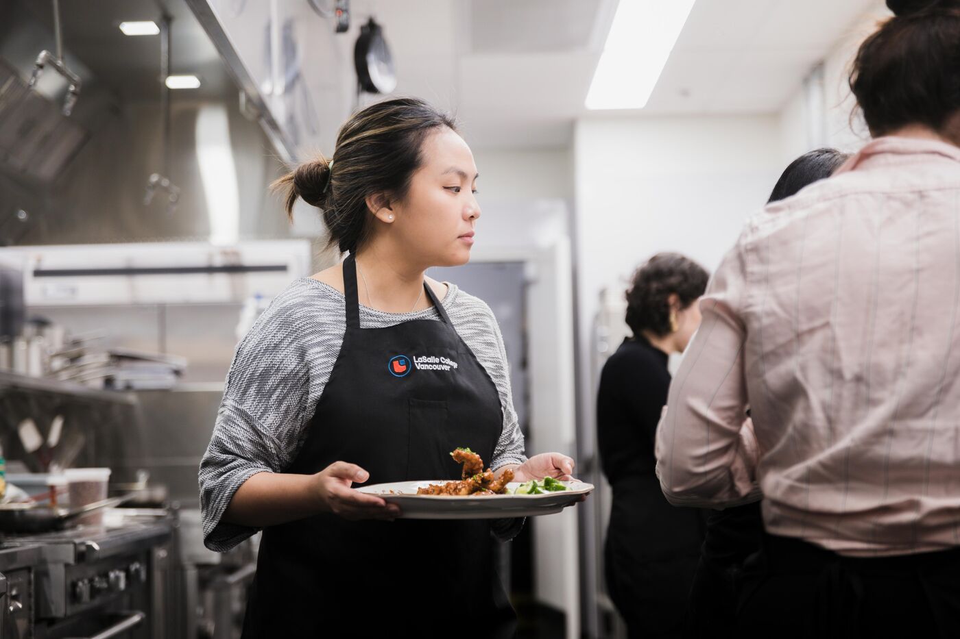 A woman wearing a black apron carries a plate of food in a professional kitchen setting, with other individuals working in the background.