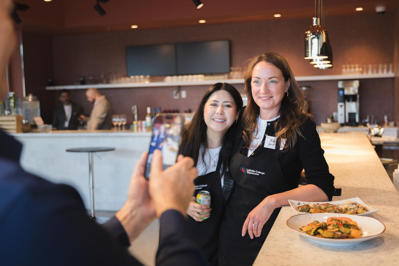 Two women smile as a third person photographs them with a smartphone in a bar or restaurant setting with a counter in front.