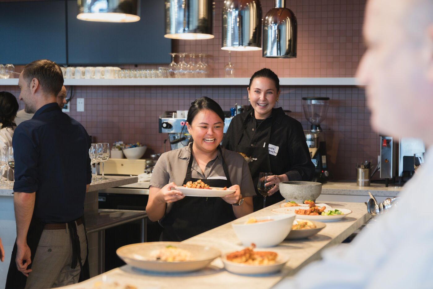Two women smile as they present plates of food in a professional kitchen setting, with other people in the background.