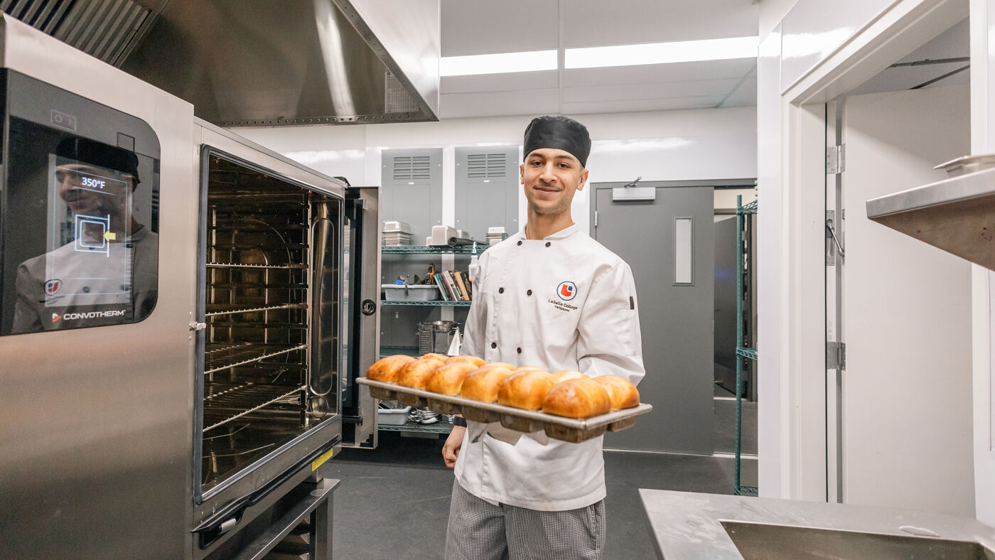 A young, smiling chef proudly presents a tray of freshly baked bread in front of an industrial oven, showcasing his culinary creation.