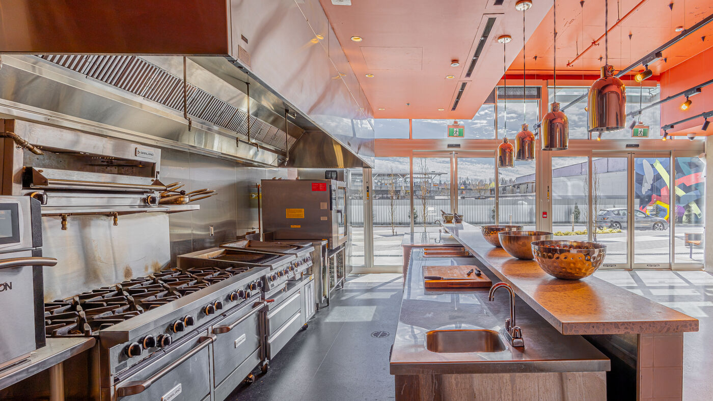 This is a professional kitchen featuring industrial stoves, a large exhaust hood, and an island with bowls. Orange lighting illuminates the space.