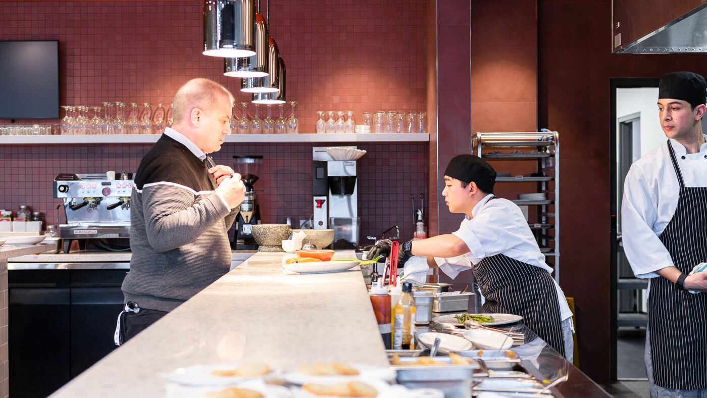 A man in a grey sweater stands at a counter while a chef in a black apron prepares food. Another chef in a white uniform stands to the right.