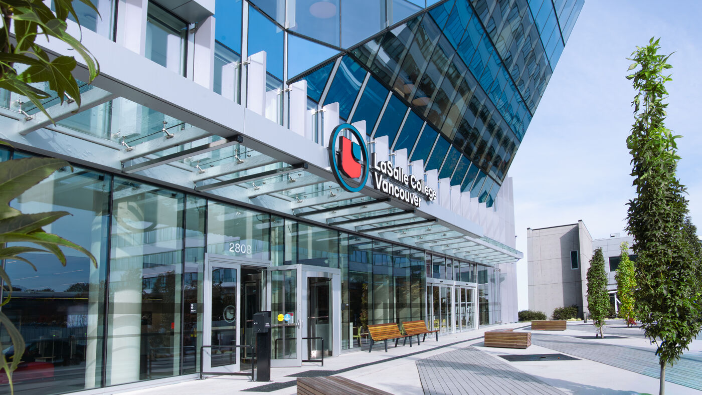 The image shows a modern building with a glass facade and a prominent logo that reads "LaSalle College Vancouver". The building has a sleek, contemporary design with a canopy over the entrance. There are benches and greenery in front of the building.