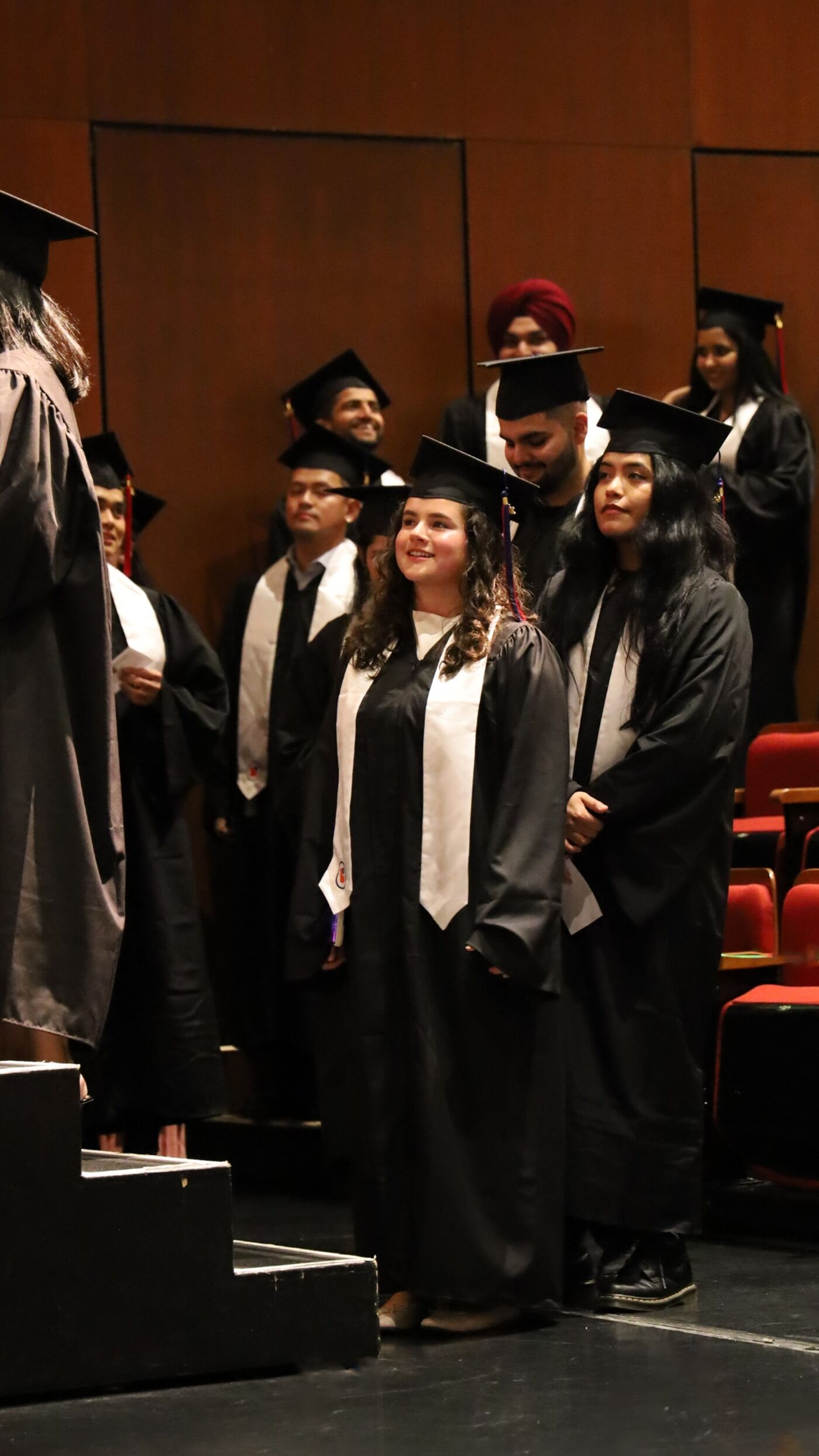 The image shows a group of people, including a woman in a cap and gown, gathered in a room, possibly a classroom or auditorium, for a graduation ceremony or event.