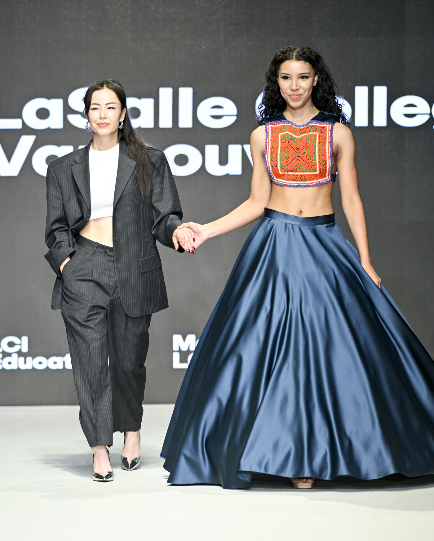 The image features a woman wearing a blue dress and a man wearing a suit, both walking down a runway or stage.