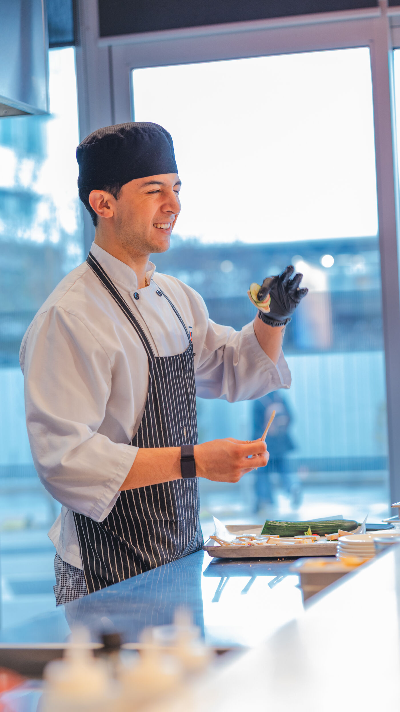 A smiling chef wearing a black hat and apron meticulously prepares sushi, showcasing a finished roll with a gloved finger.