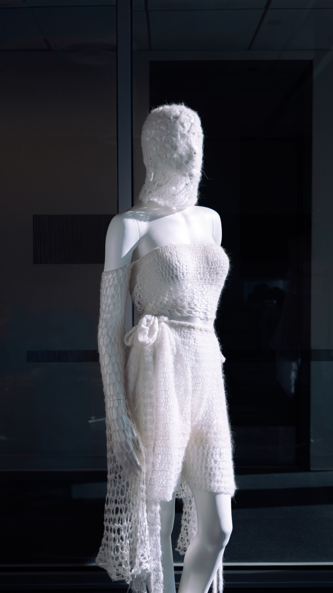 A female mannequin figure dressed in a white, crocheted outfit with long, flowing sleeves, standing in front of a dark window.