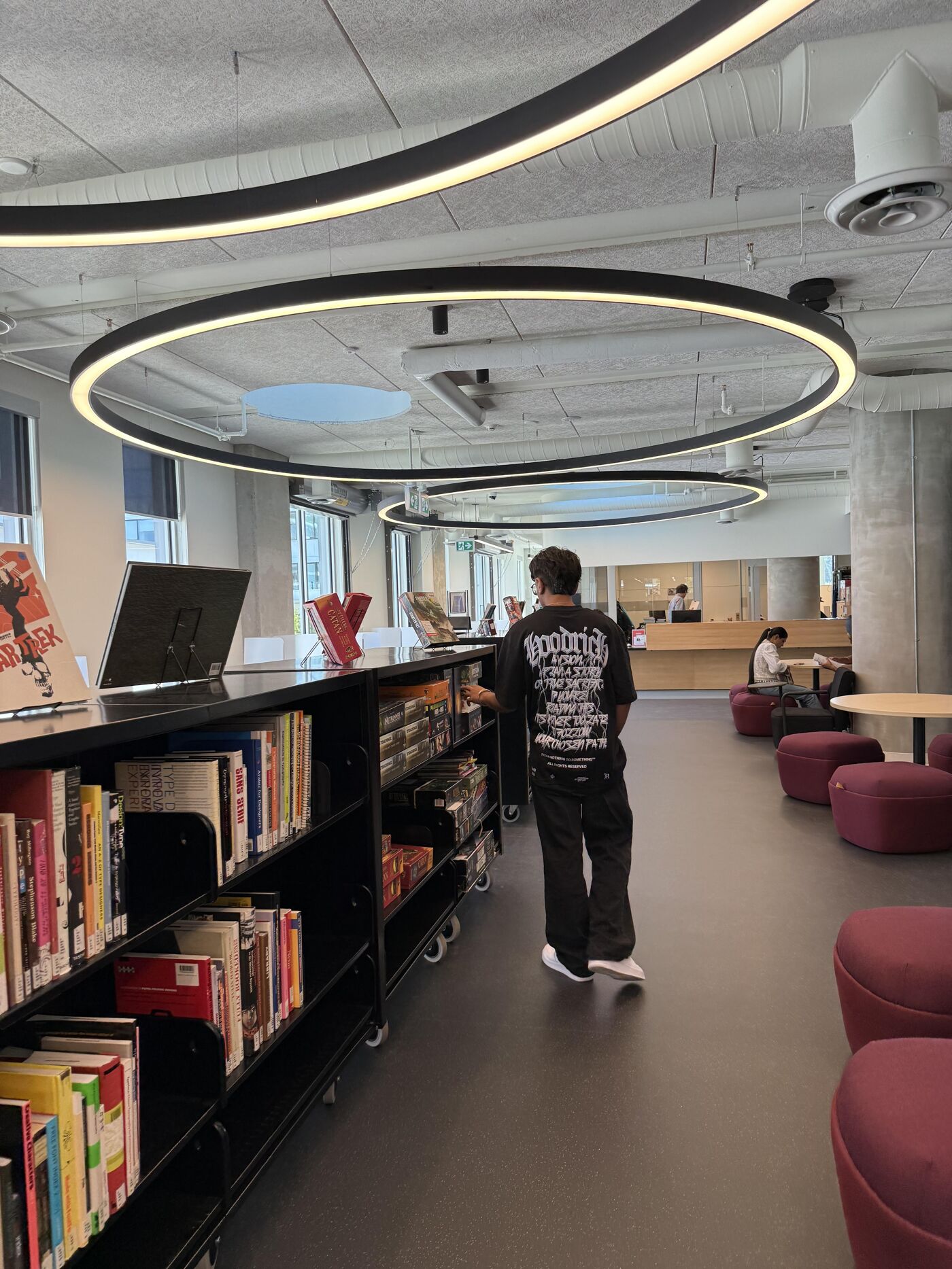 The image shows a person standing in a library or bookstore. There are bookshelves filled with books on the left, and circular light fixtures hanging from the ceiling. There are also some chairs and tables in the background.