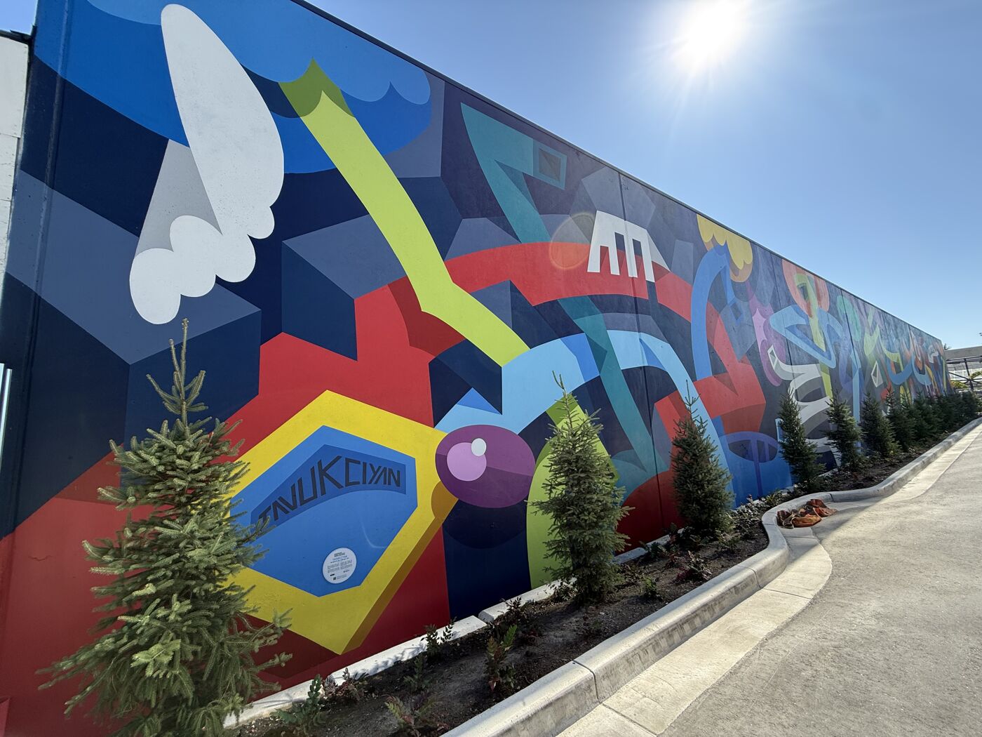 The image shows a colorful mural on the side of a building, featuring abstract shapes and patterns in various colors like blue, red, yellow, and green. Small trees are planted along the base of the wall.