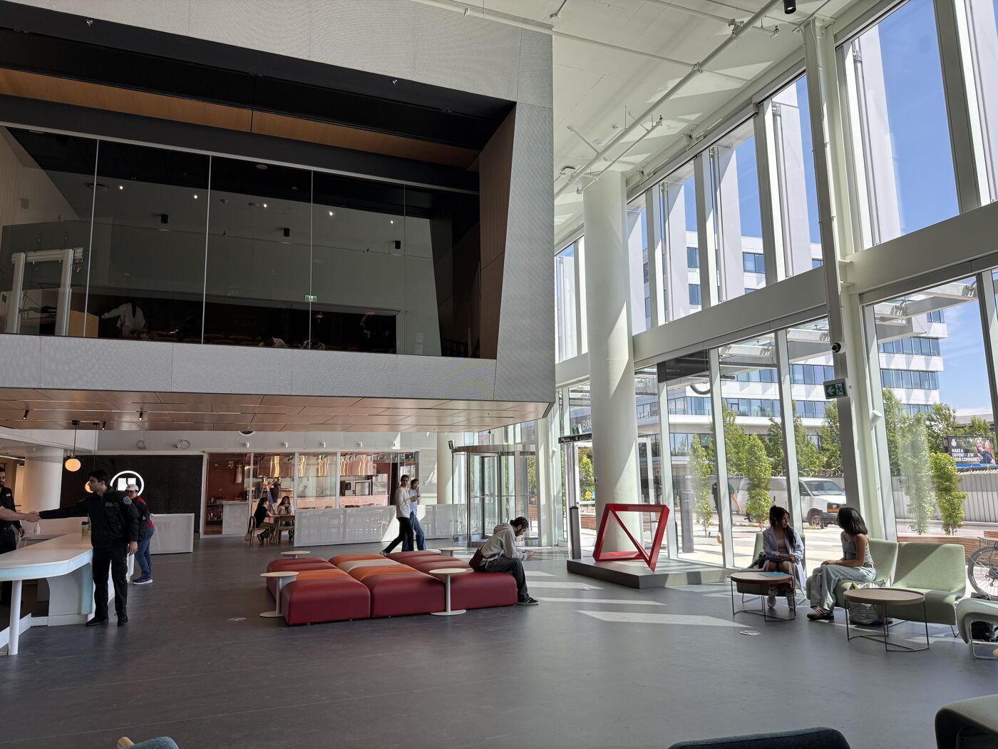 The image shows a spacious and modern lobby with large windows, allowing ample natural light. There are seating areas with red couches and chairs, and people are seen relaxing and working in the space. The architecture features clean lines and a minimalist design.