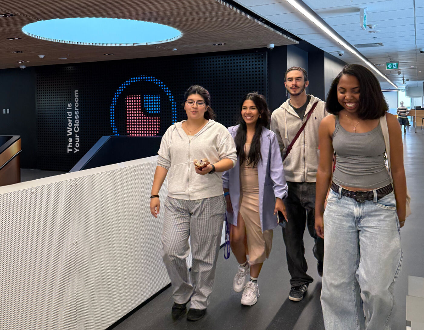 The image shows four people walking in a modern building. Three women and one man are walking side-by-side, smiling. The background features a wall with a digital display and architectural lighting.