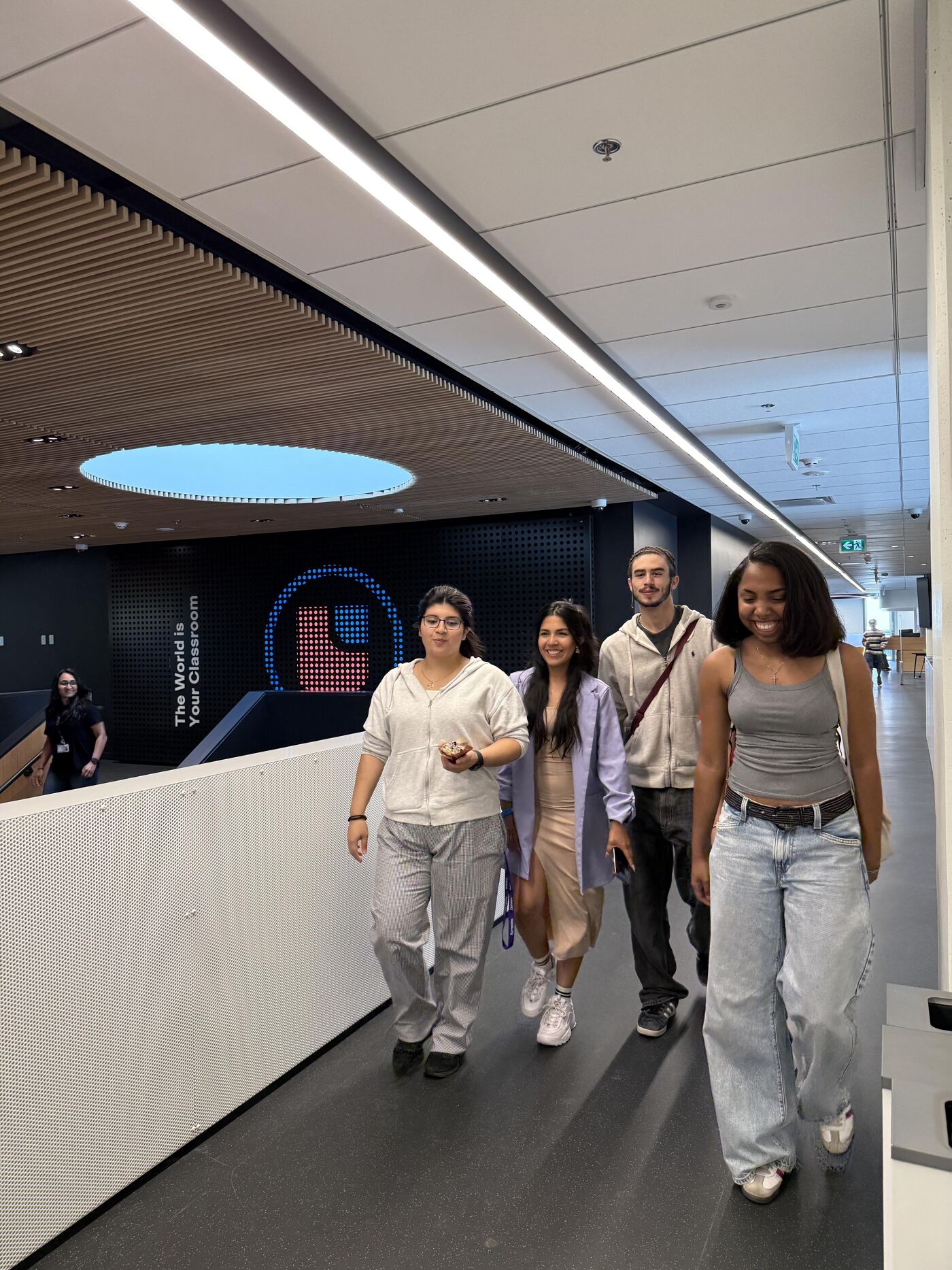 In an indoor setting, four individuals are walking along a corridor. The group consists of three women and one man, all casually dressed. The corridor features modern architecture with a combination of natural and artificial lighting.