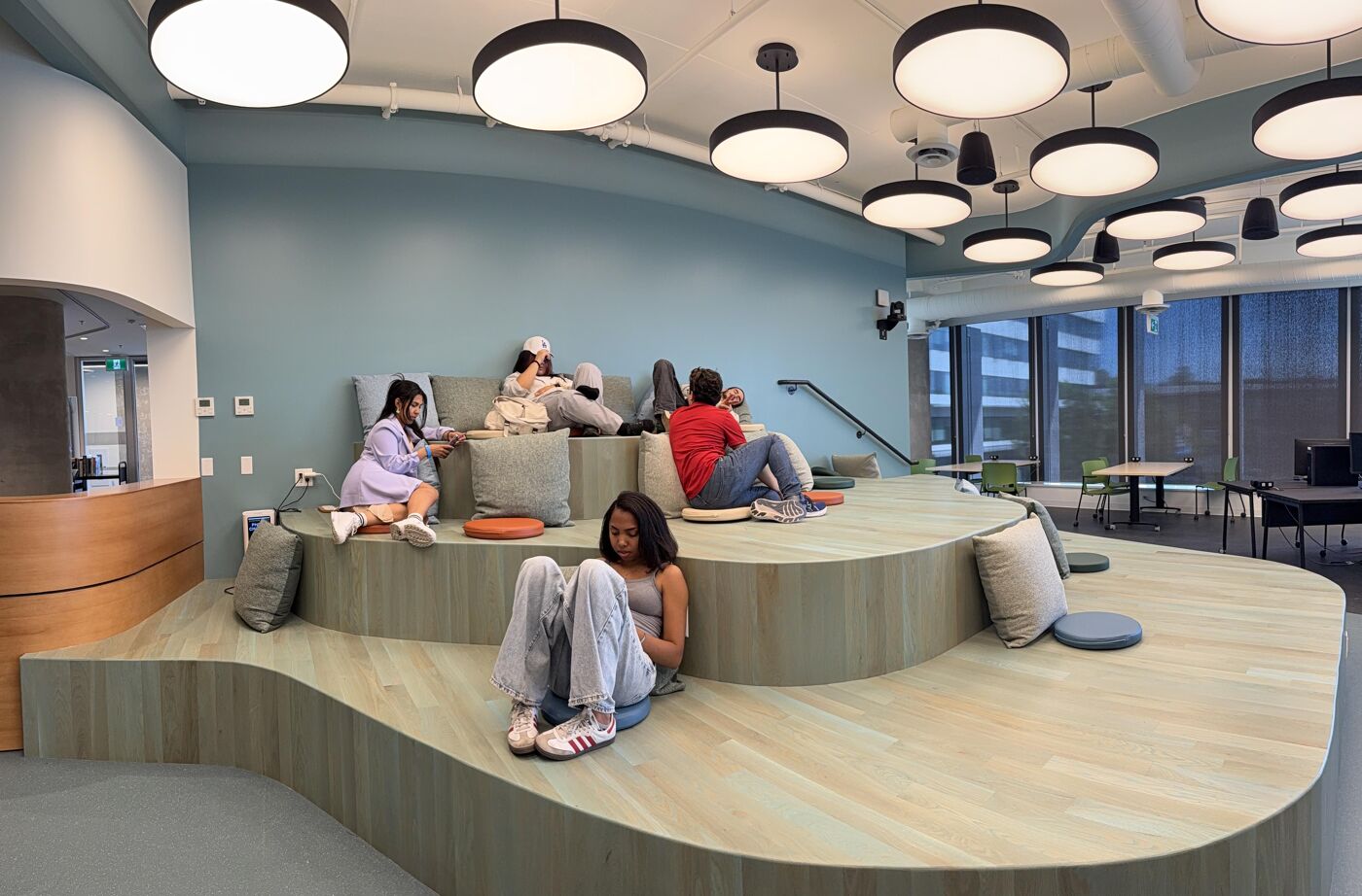 The image shows a modern, multi-tiered seating area with several people relaxing and studying. The space features light wood flooring, soft cushions, and natural light streaming through large windows, creating a comfortable and inviting atmosphere.
