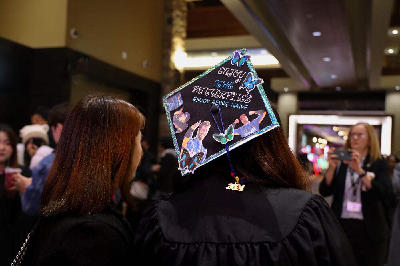  A graduate wears a customized cap featuring colorful butterflies, glittery accents, and the phrase "Enjoy the butterflies, enjoy being naïve." The festive atmosphere includes celebratory attendees and photography capturing the moment.