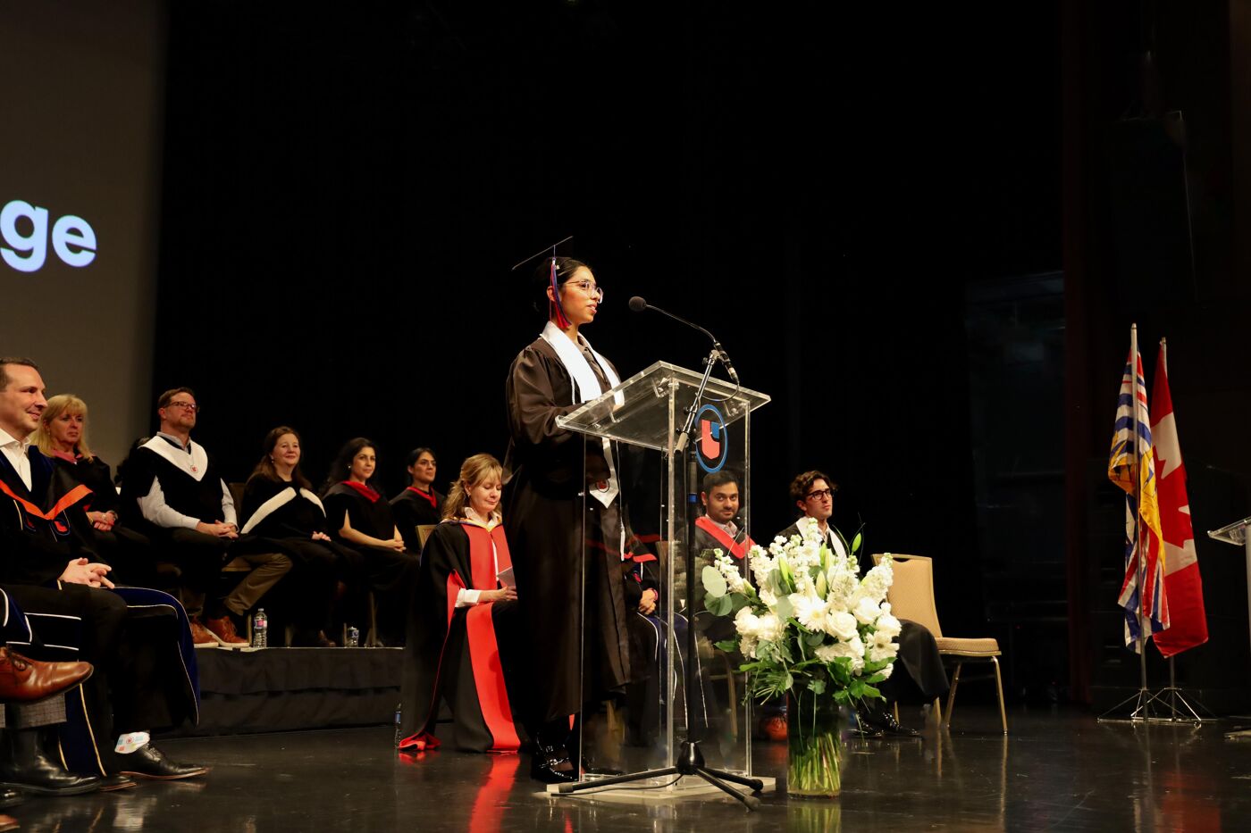 A graduate in cap and gown delivers a speech at a podium during a formal graduation ceremony. Faculty members in academic regalia sit on stage, with floral arrangements and flags adding to the dignified atmosphere.