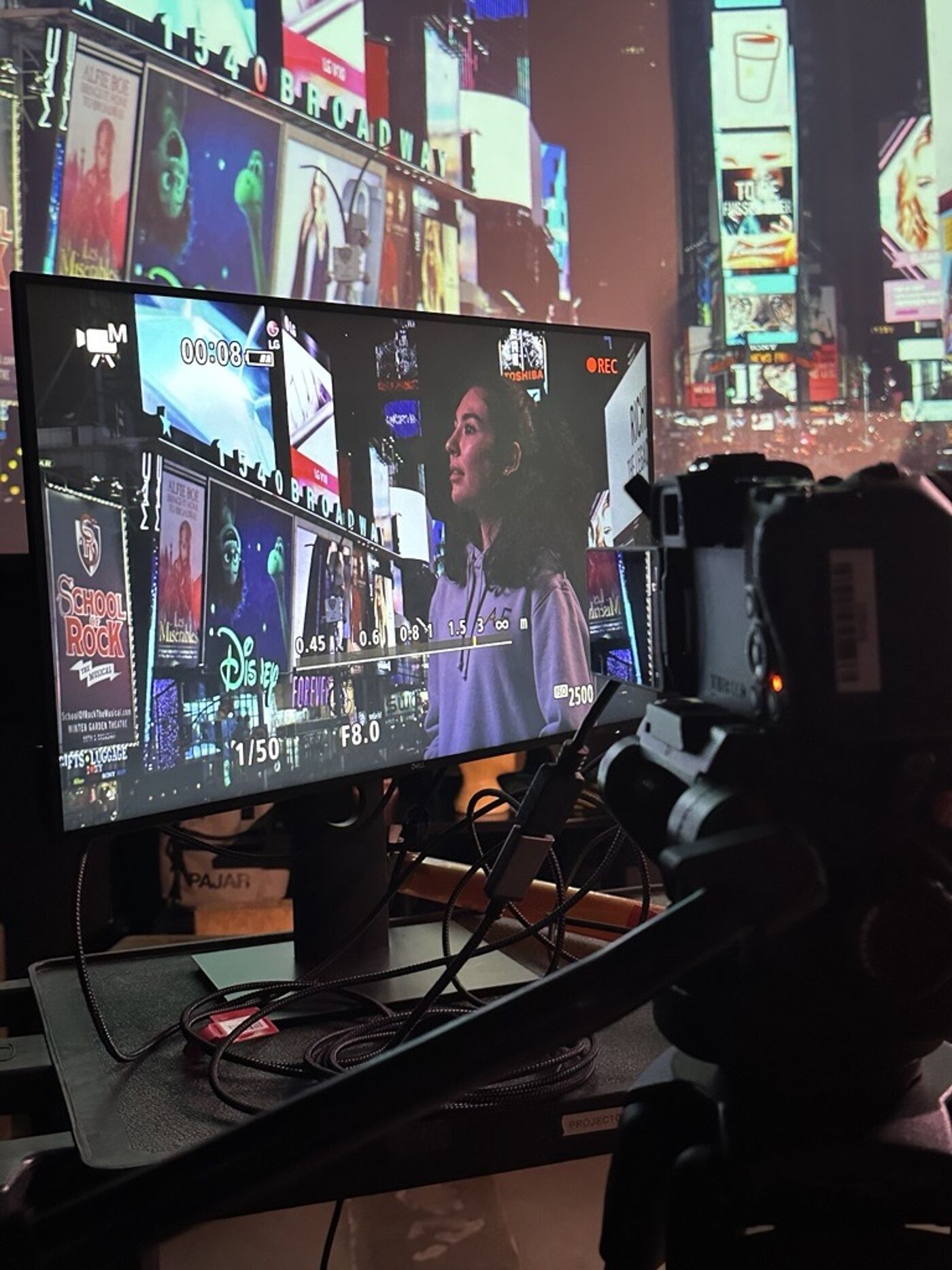 A monitor displays a scene being filmed, featuring a woman against a backdrop of bright urban billboards, likely a projection of Times Square. The setup includes a camera and cables, emphasizing a professional production environment.