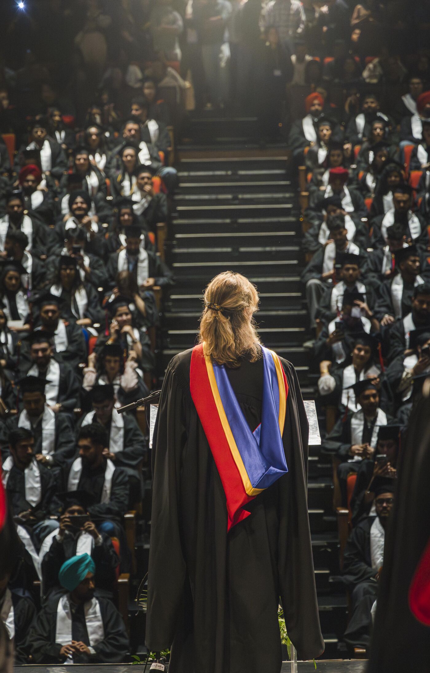 A woman in academic regalia stands facing a large audience of graduates seated in rows. The graduates are wearing black gowns and some have head coverings. A staircase leads up to the higher rows of seating.