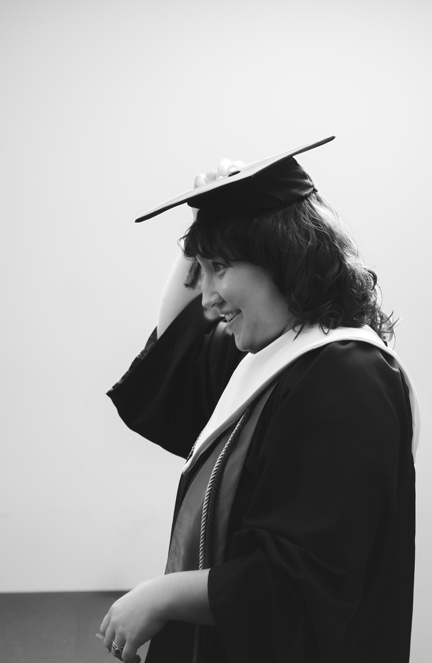 The black and white image shows a woman in graduation attire, including a gown and mortarboard hat. She is smiling and appears to be adjusting her hat. The background is a plain white wall.