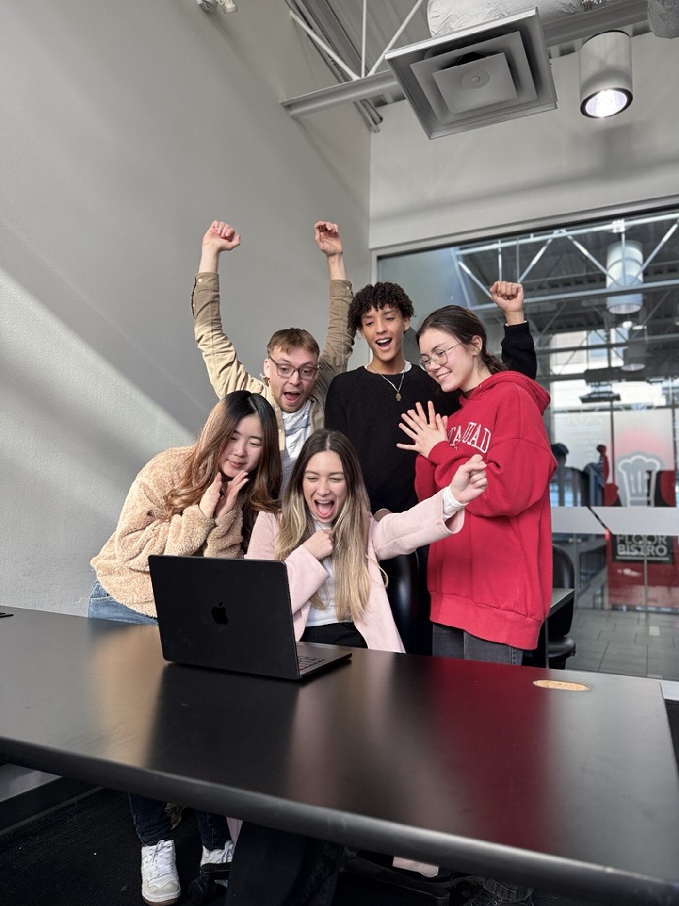 Five young adults, three women and two men, are gathered around a laptop, two of them with their arms raised in celebration.