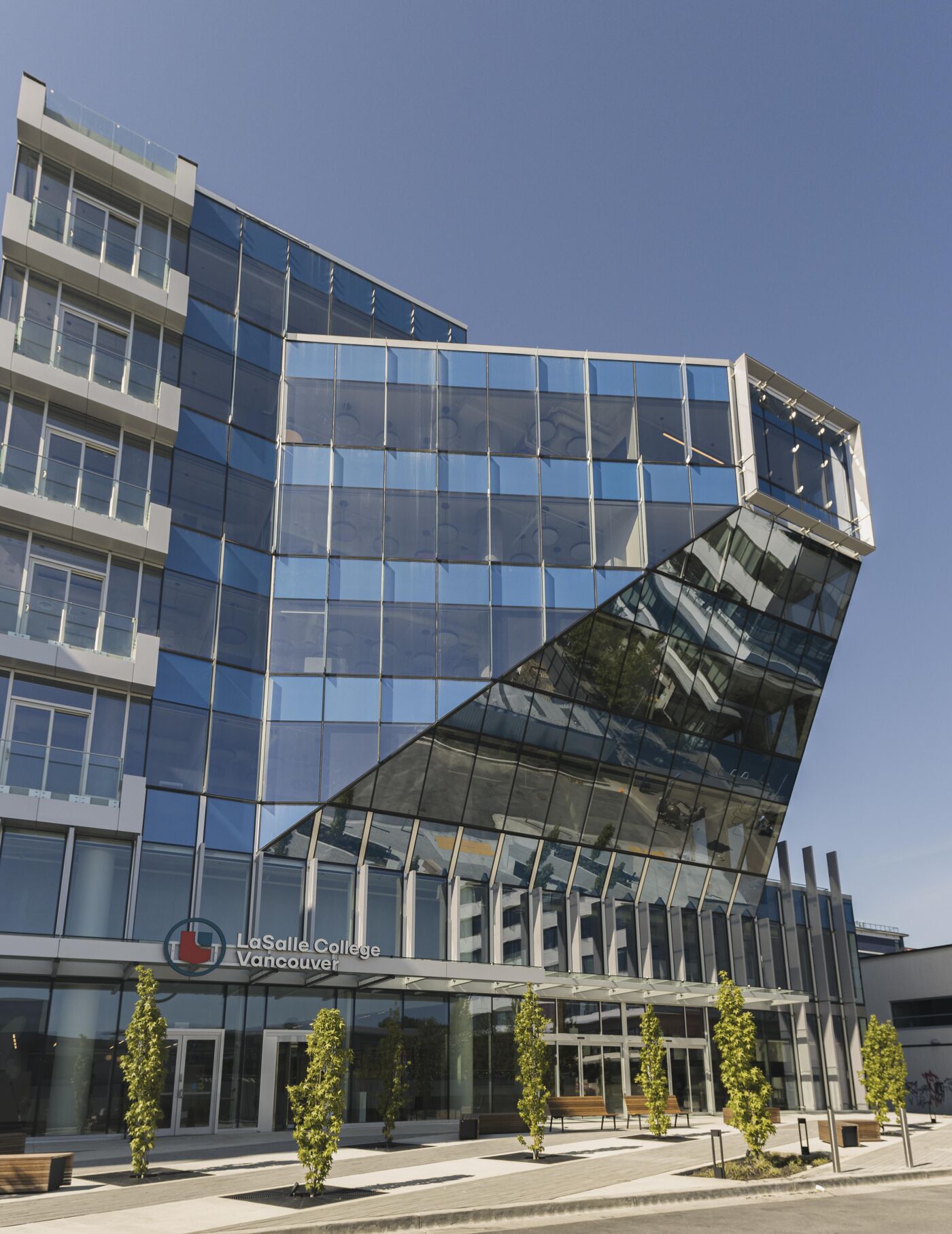 The image shows a modern, angular building with a glass facade, reflecting the sky. The building features a unique design with a prominent diagonal section. Trees line the entrance, adding a touch of greenery to the urban landscape.