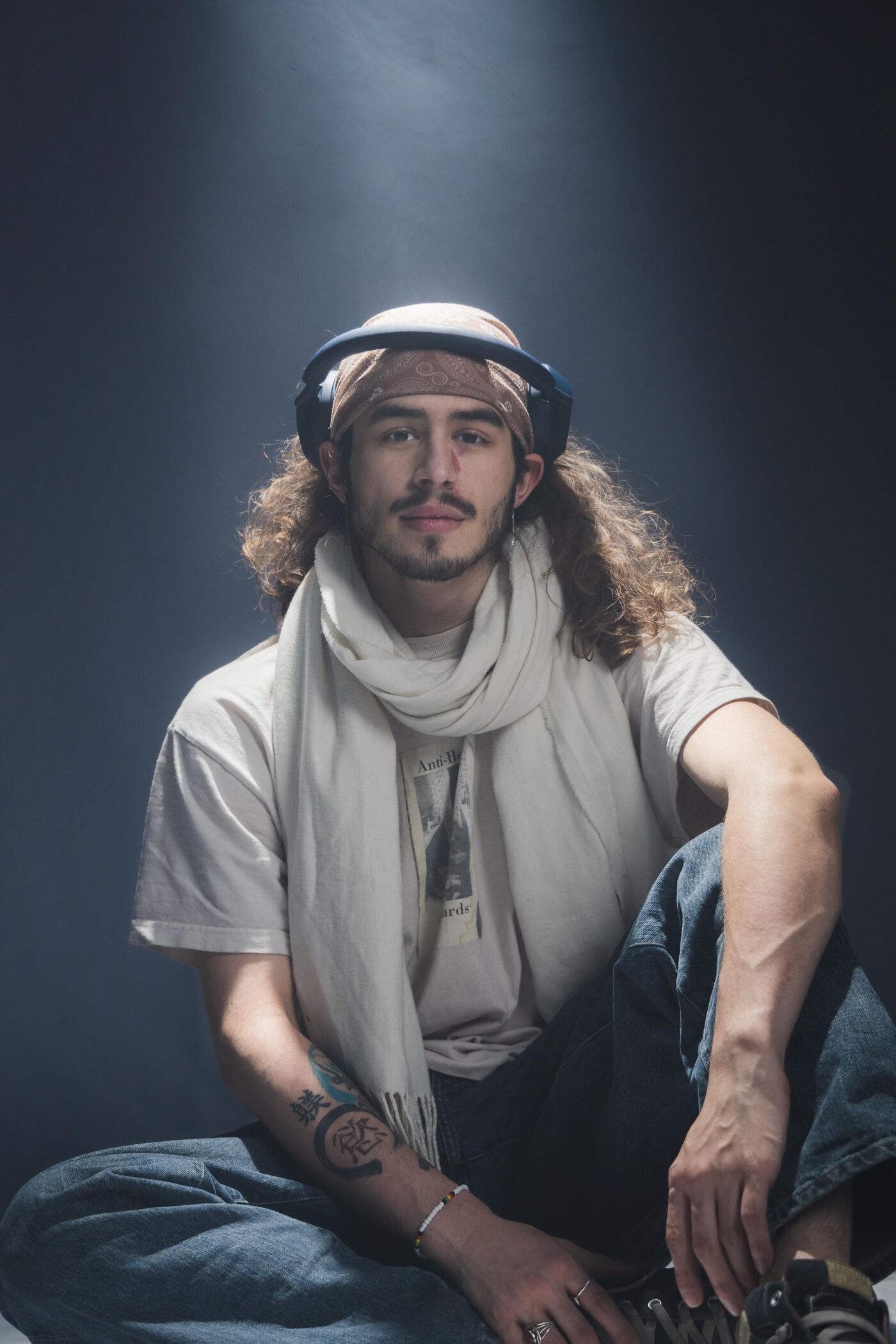 A young man with long brown hair, wearing headphones, a bandana, a white t-shirt, a light-colored scarf, and jeans, sits cross-legged on a dark background, looking directly at the camera.