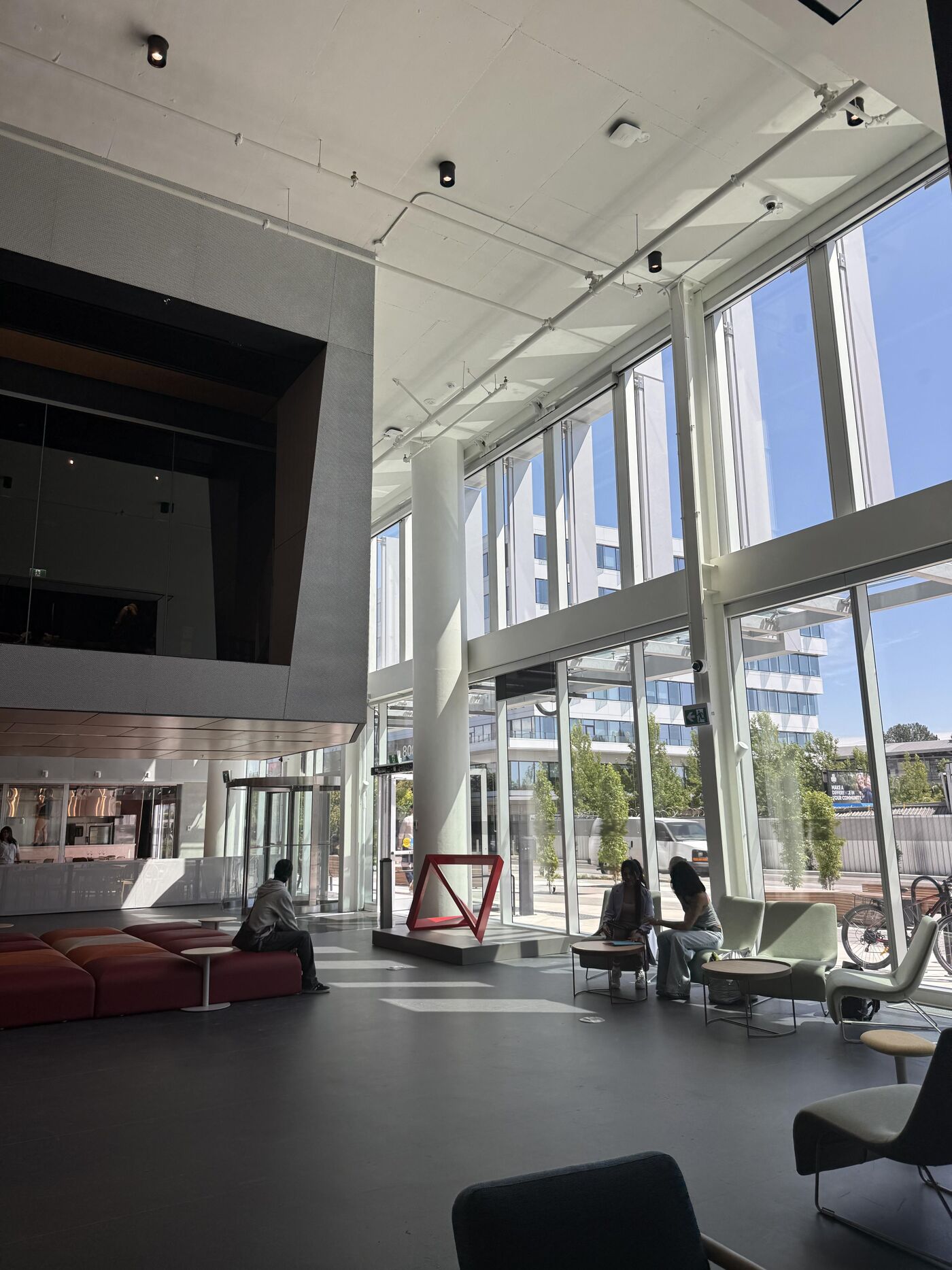 The image shows a spacious, modern lobby with large windows offering ample natural light and views of the outside. People are seated in the lobby, and there's a mix of contemporary furniture and architectural elements.