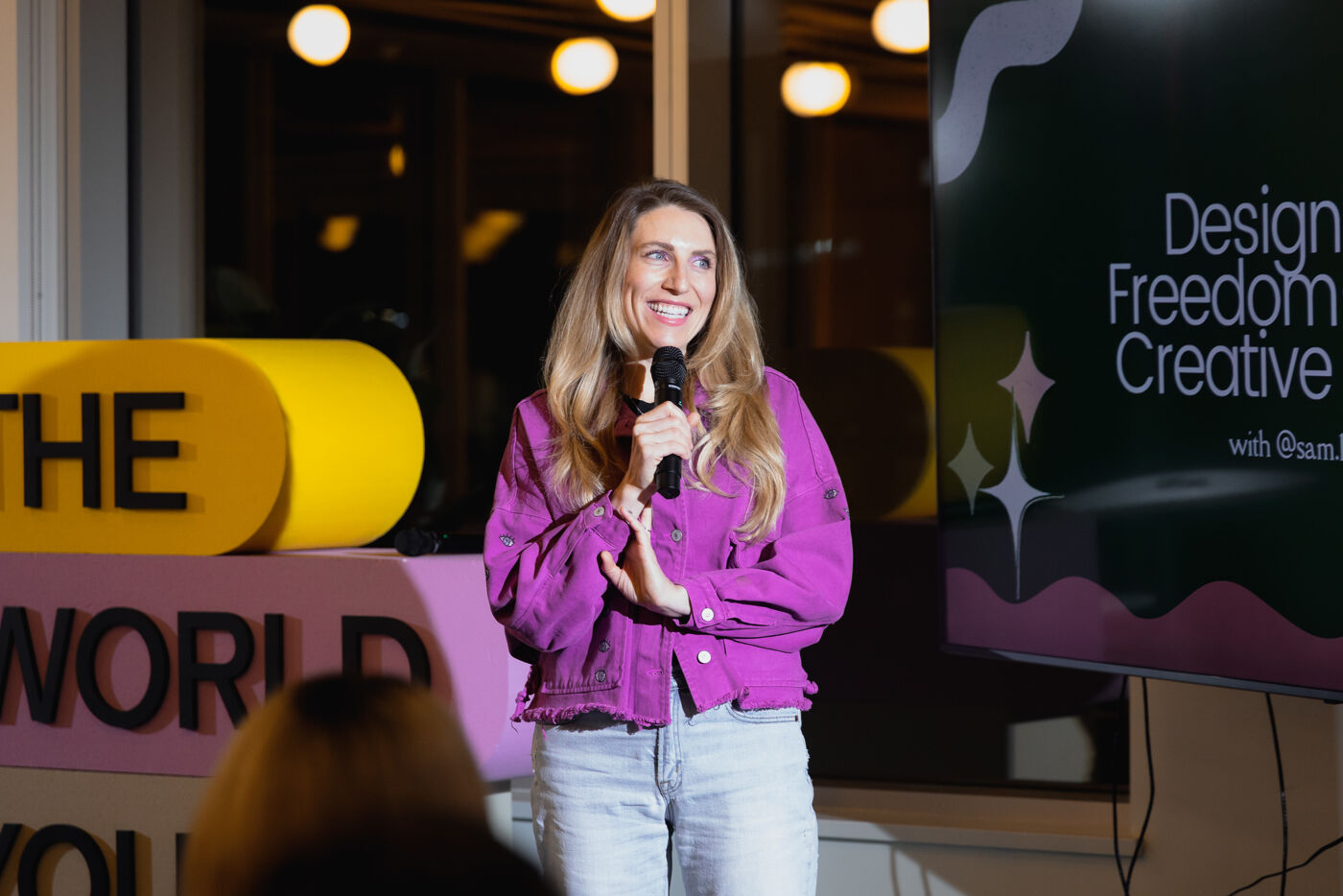A woman with blonde hair is speaking into a microphone. She is wearing a purple jacket and jeans. Behind her is a screen with text.