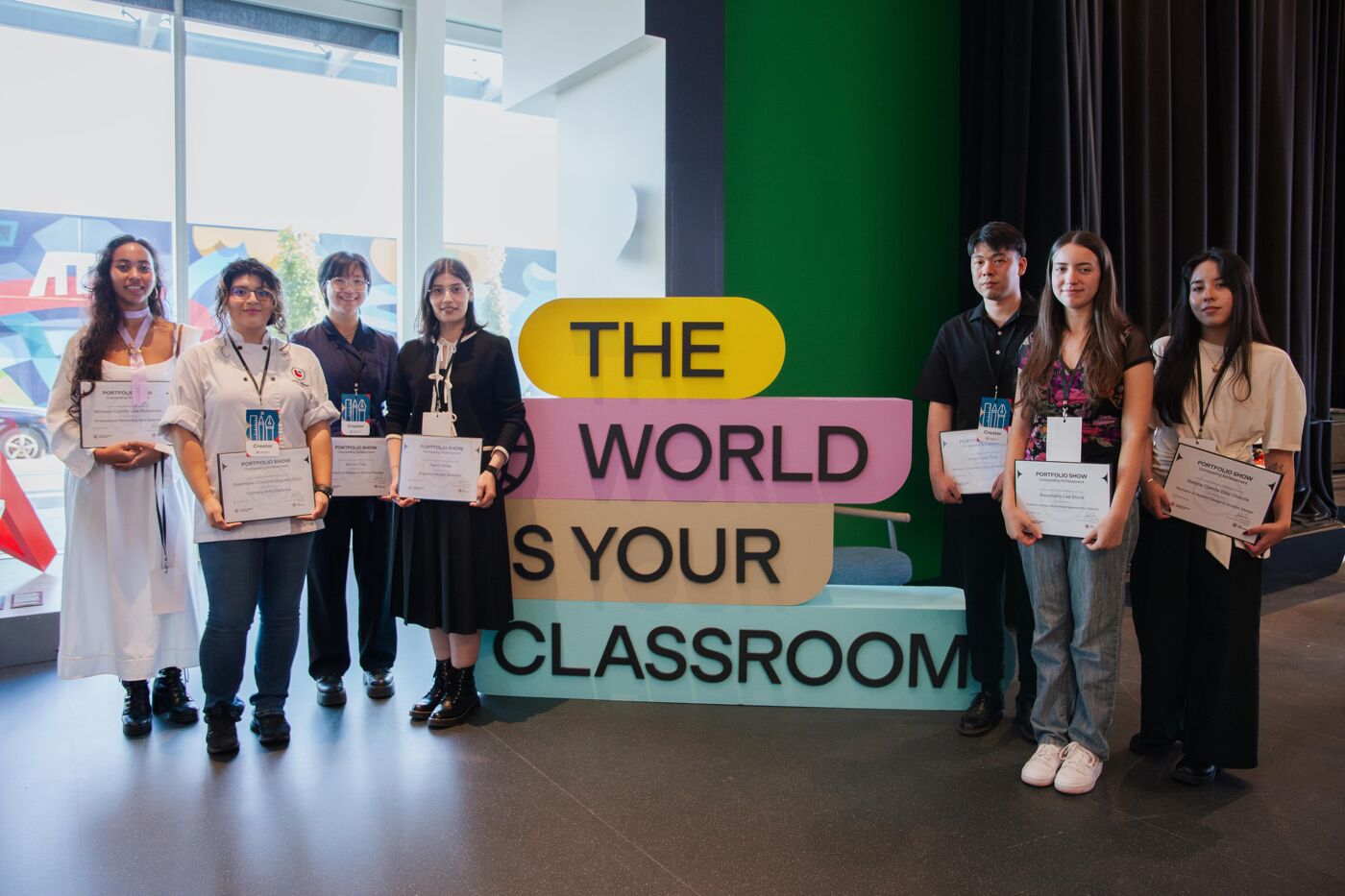 The image shows a group of eight people standing in front of a sign that reads "The World Is Your Classroom." They appear to be at an event, possibly an awards ceremony, as some are holding certificates.