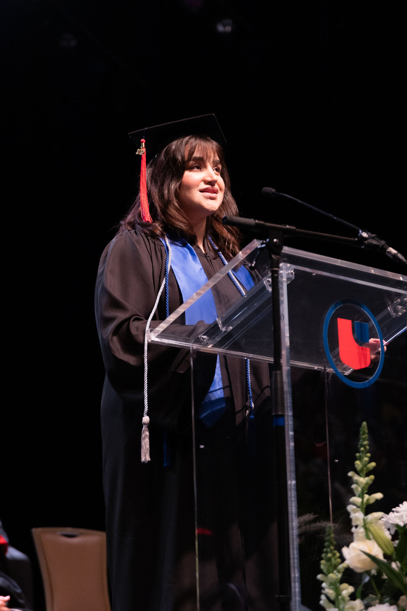 In the image, a graduate in a cap and gown is giving a speech at a podium. She appears to be at a graduation ceremony, addressing an audience.