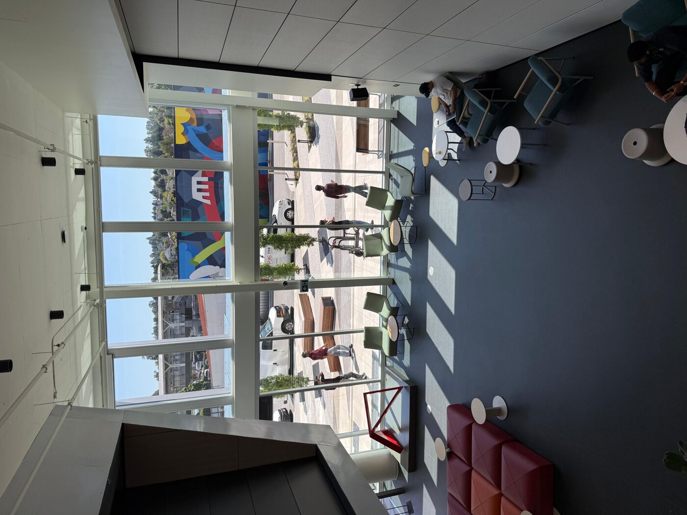 The image shows a spacious, modern interior with large windows overlooking an outdoor area. There are various seating arrangements, including couches and chairs, creating a relaxed and open atmosphere.