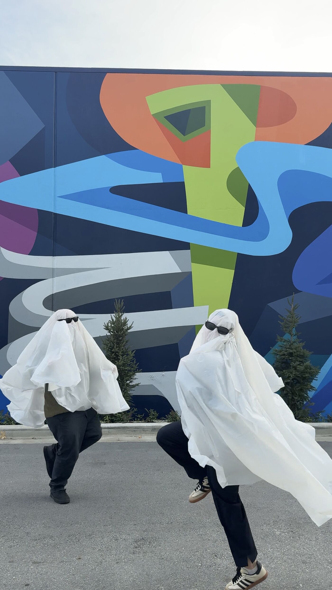 Two people dressed as ghosts wearing sunglasses are posing in front of a colorful mural. They are both wearing white sheets and dark pants, creating a playful and spooky scene.