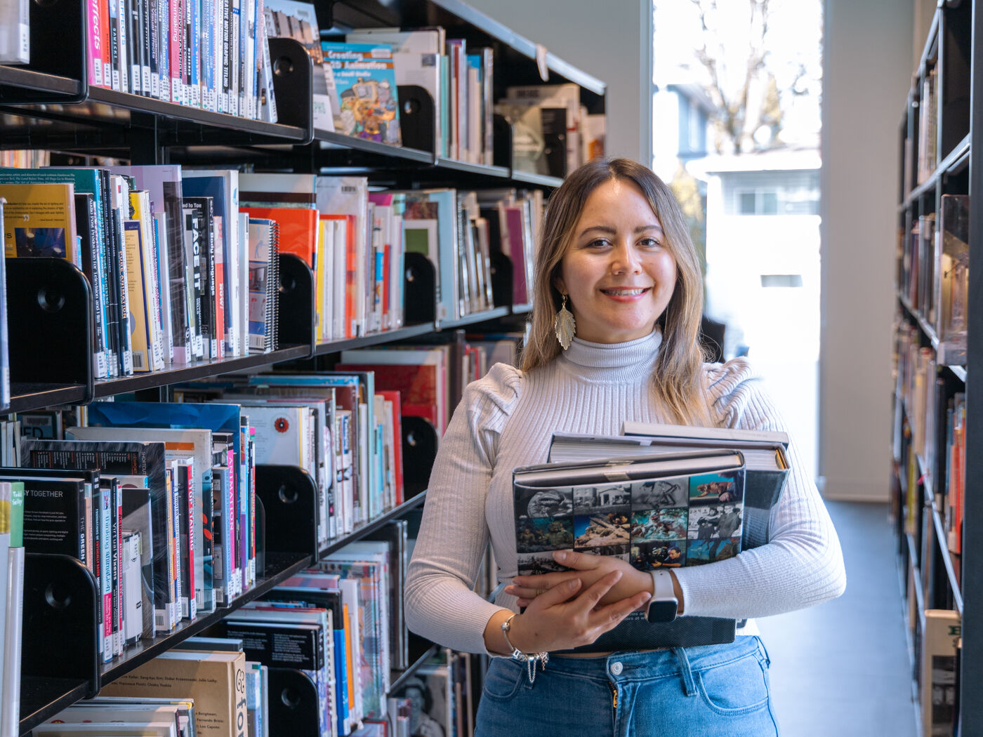 A smiling woman stands in a library, holding books. She is surrounded by bookshelves filled with books, suggesting a love for reading and learning.