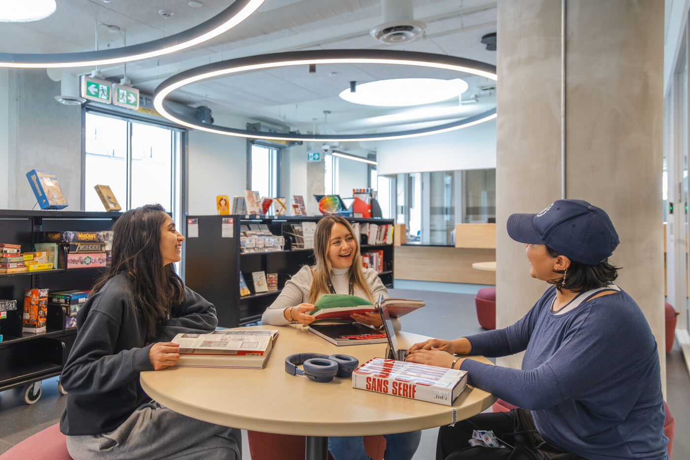 Three women are sitting around a table in a library, engaged in conversation and looking at books. The library has a modern design with bookshelves and circular lights.
