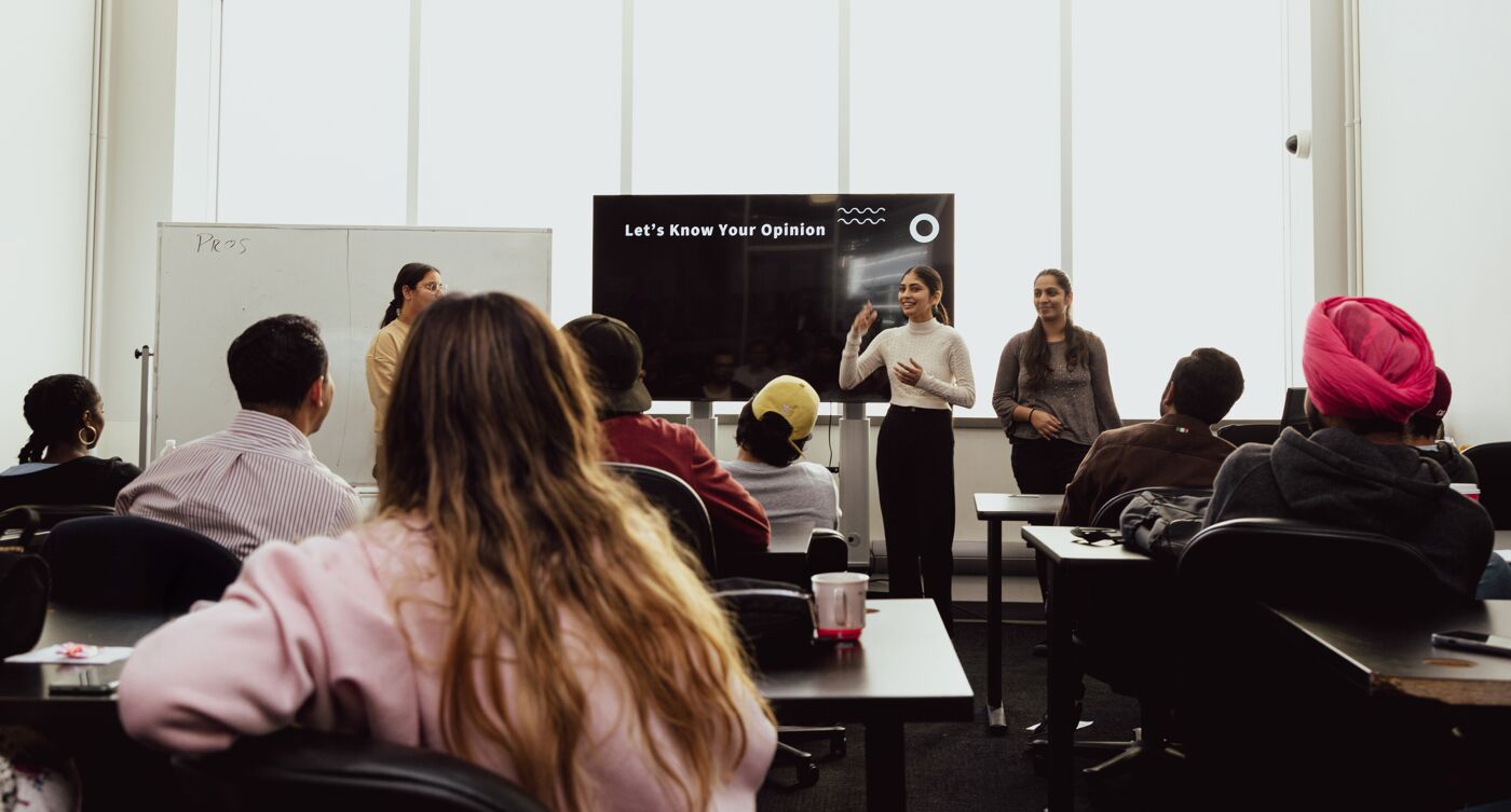 Students attentively listen to a presentation, as the speaker invites audience participation in a modern classroom setting.