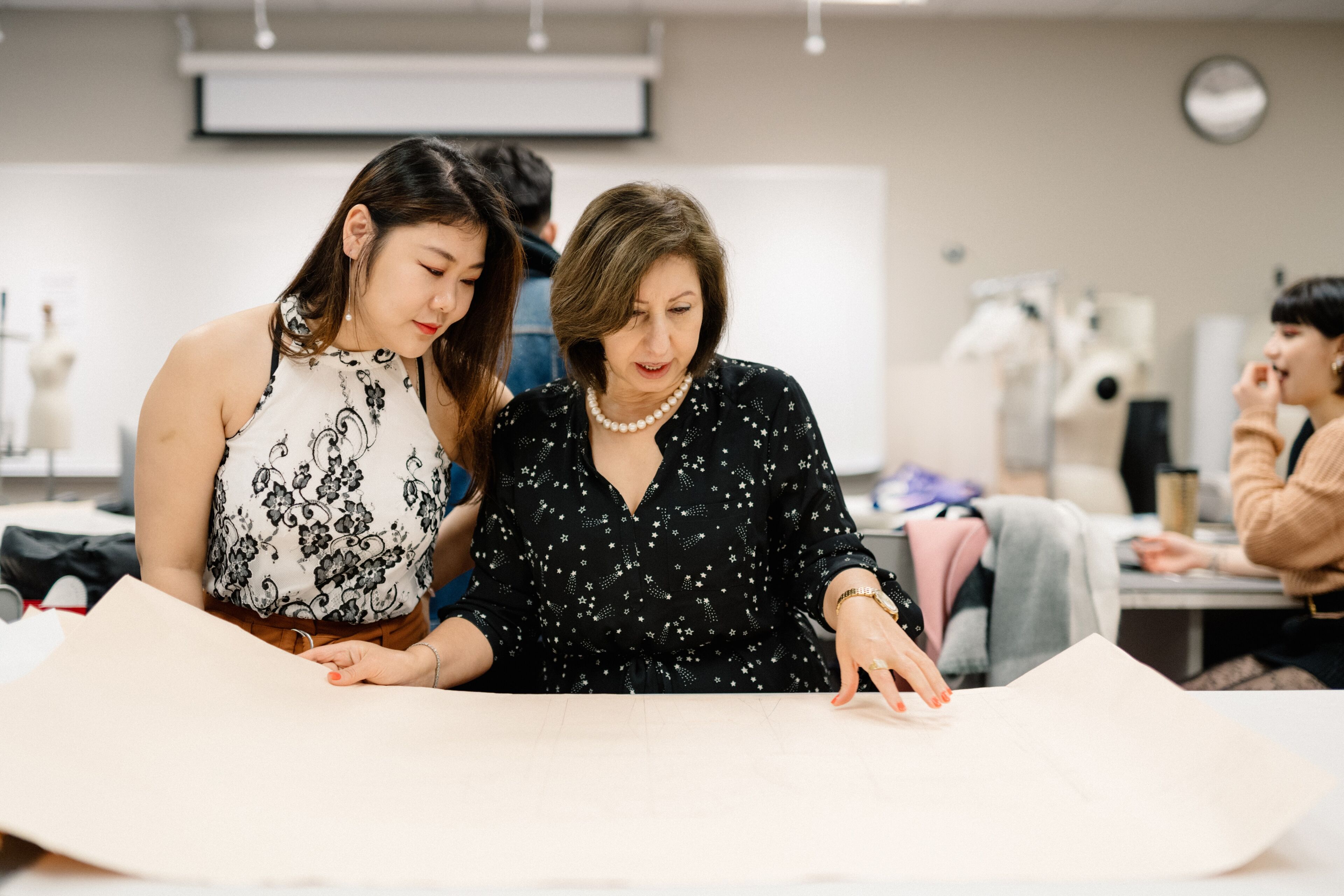 An experienced designer guides a young student through pattern making in a fashion studio.