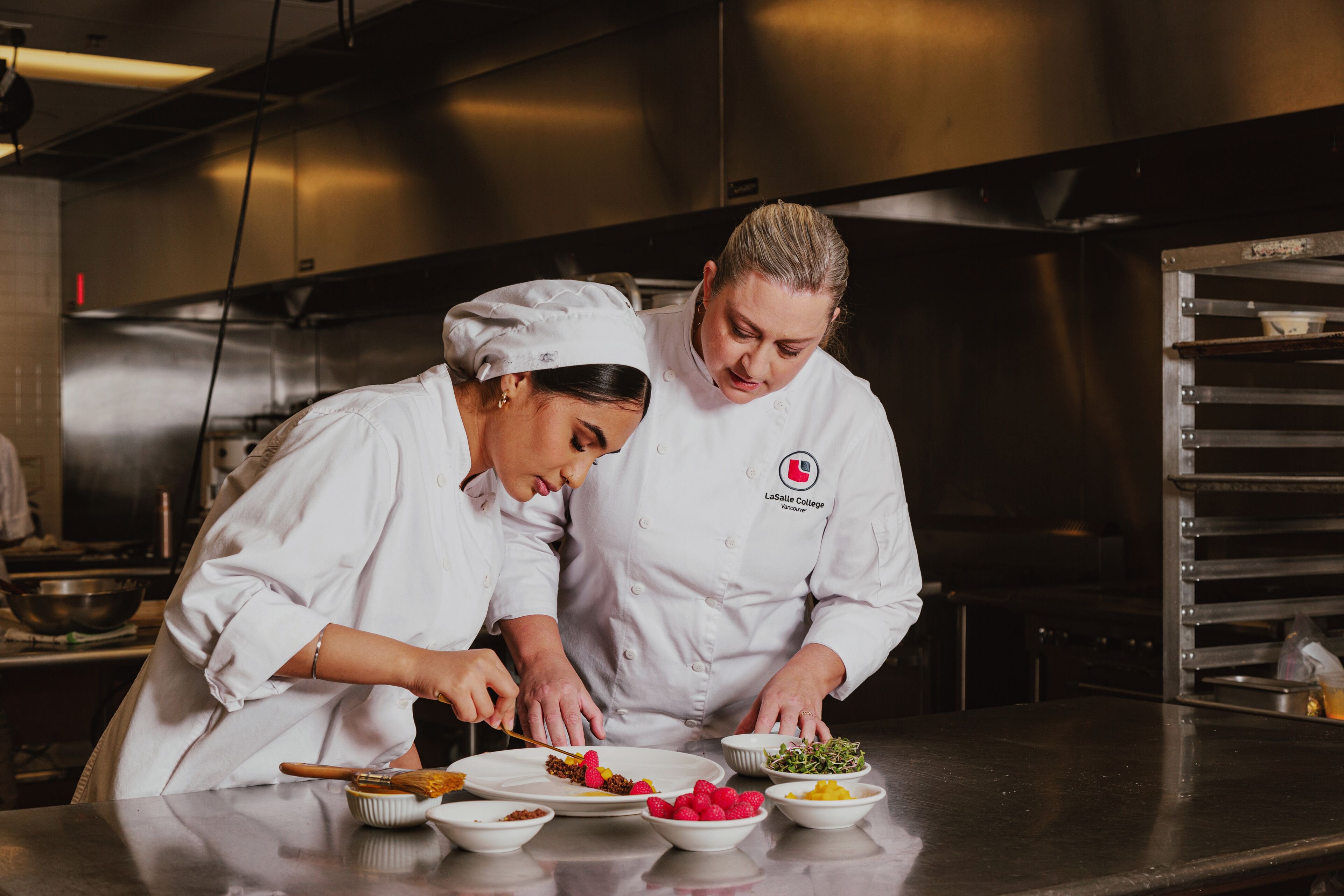 A seasoned chef guides a young cook in the art of plating, combining expertise with fresh talent.