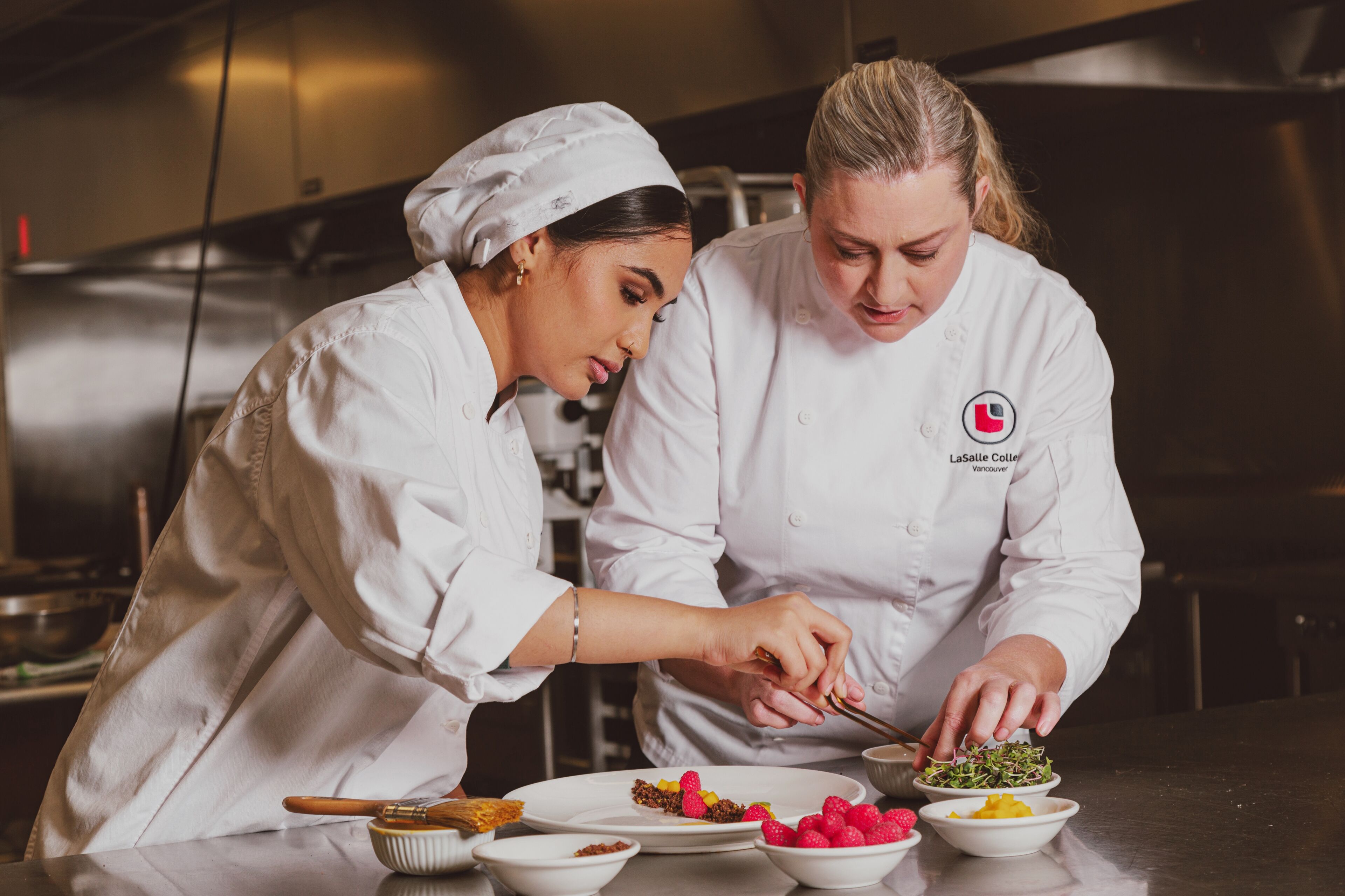 A seasoned chef guides a young cook in the art of plating, combining expertise with fresh talent.