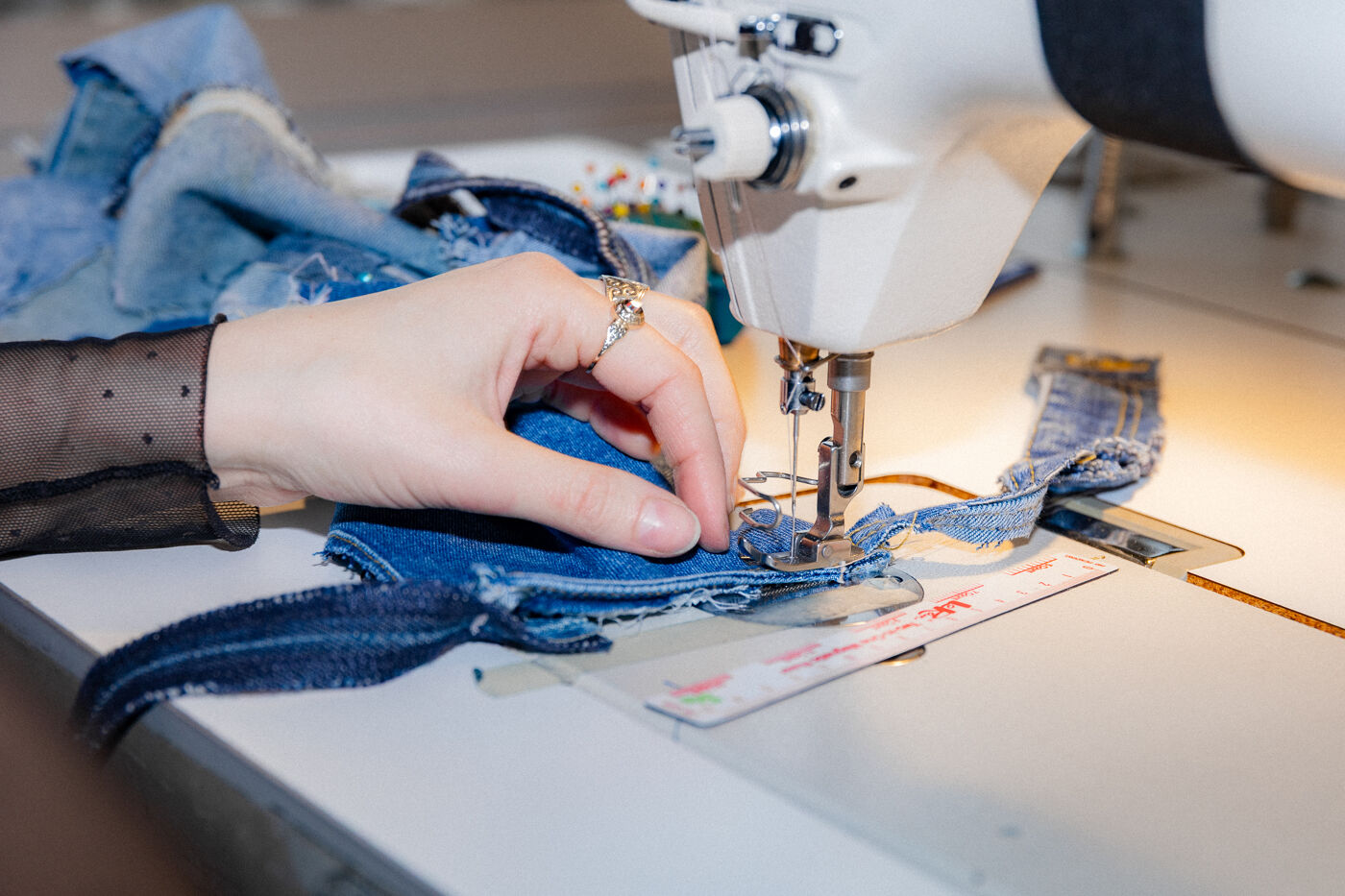 A hand with a ring is sewing denim fabric on a sewing machine. The fabric appears to be undergoing transformation or repair.