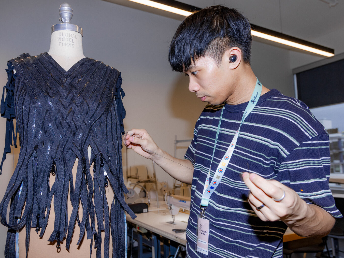 An Asian fashion designer works on a black dress with fringe hanging from a mannequin in a workshop with fluorescent lights overhead.